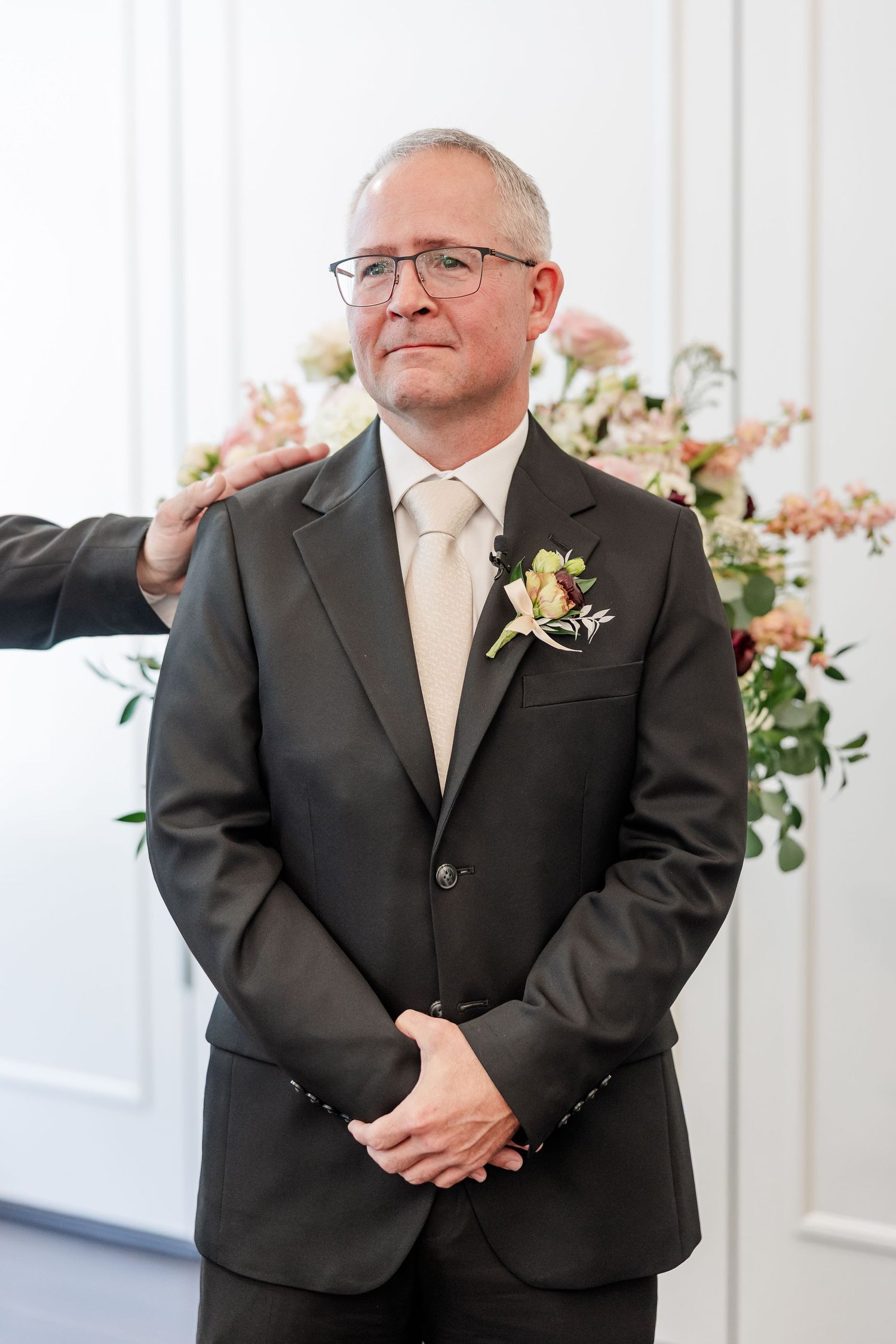 Groom in a gray suit and glasses stands with hands clasped and a floral boutonniere as a hand rests on his shoulder at a wedding ceremony.