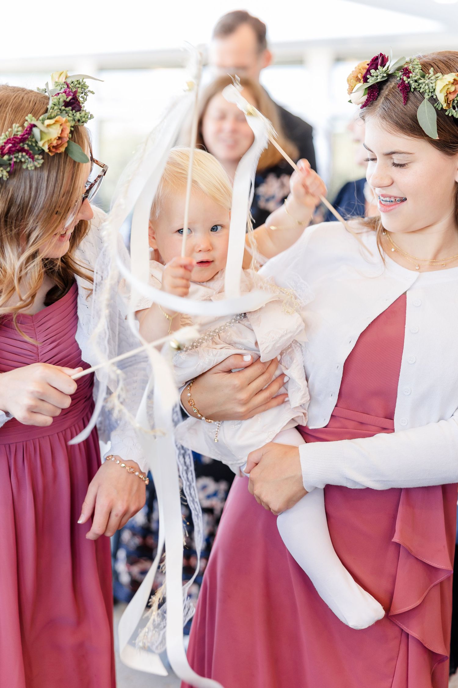 Two bridesmaids in mauve dresses and floral crowns hold a smiling baby with white ribbon wands at a wedding celebration.