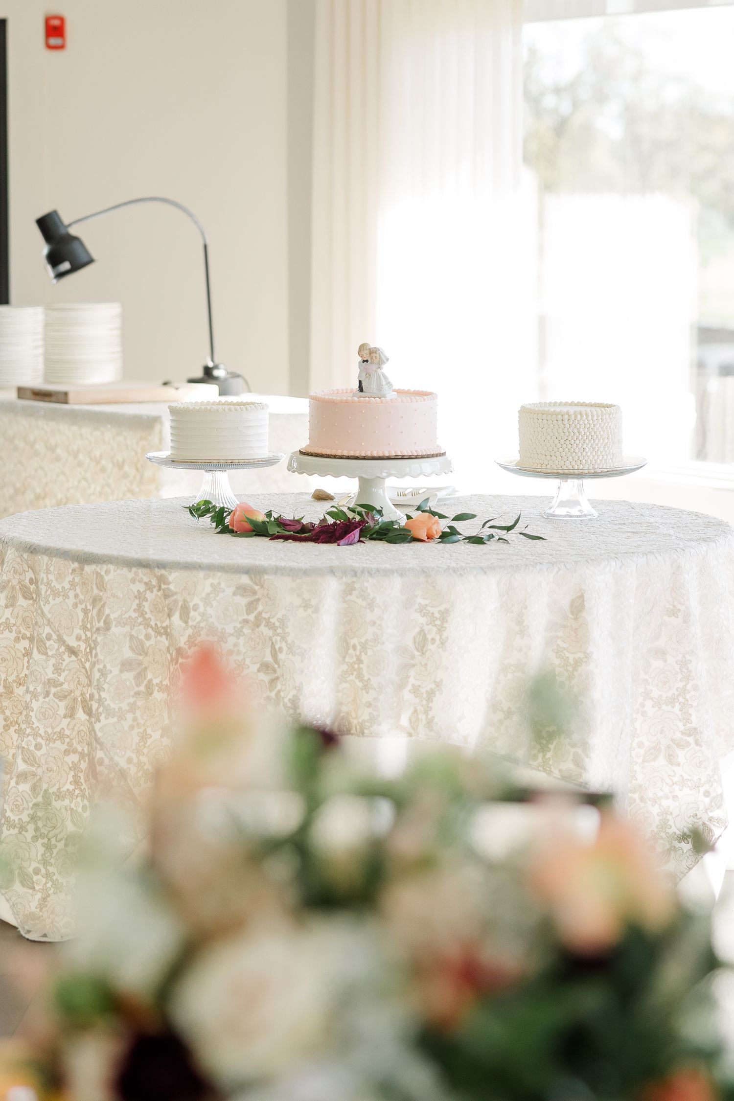 Wedding cake table with a pink cake with figurine topper and two white cakes on stands, draped in lace linen with floral garland.