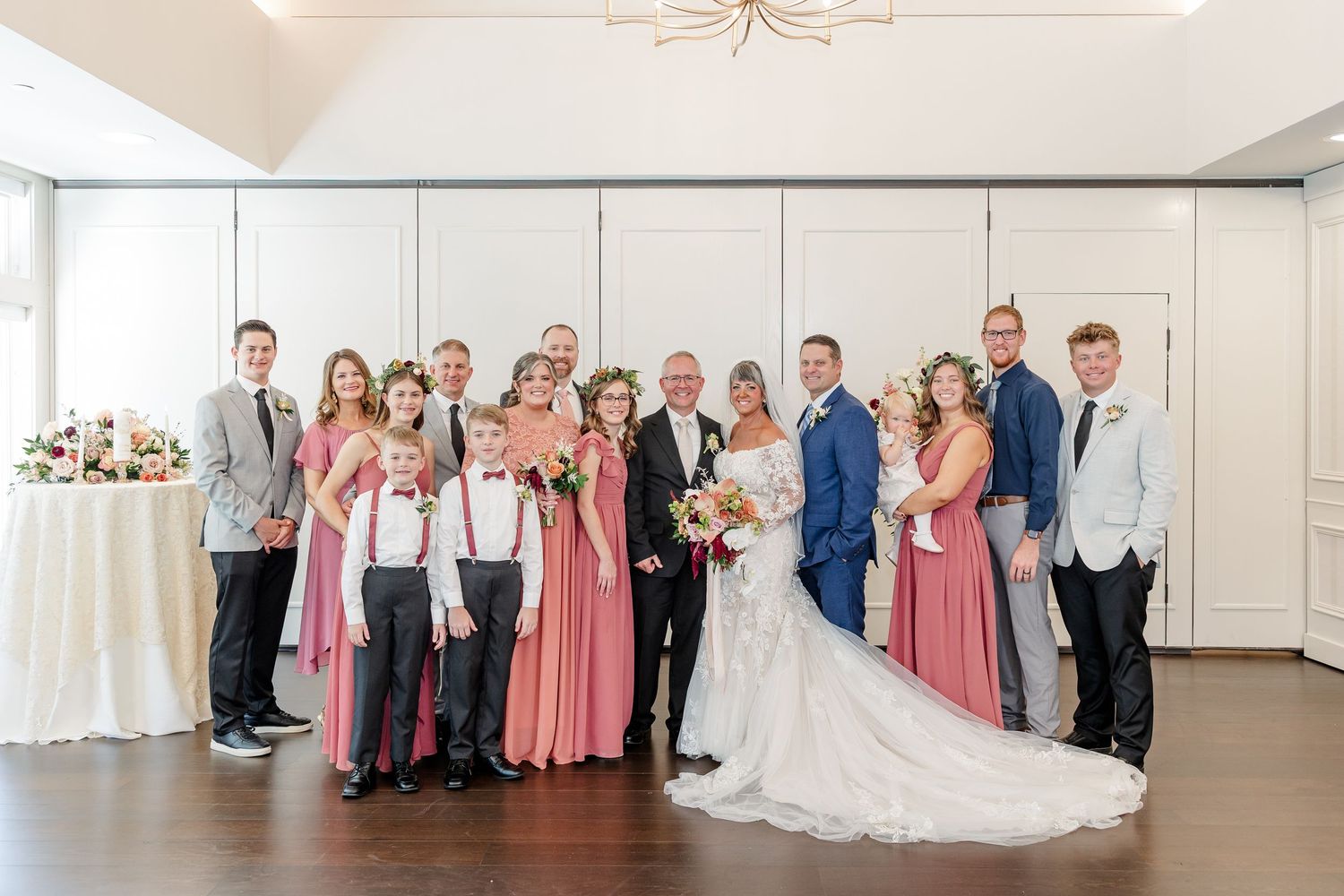Formal wedding party portrait with the bride and groom, bridesmaids in dusty rose dresses, groomsmen, and young ring bearers indoors.