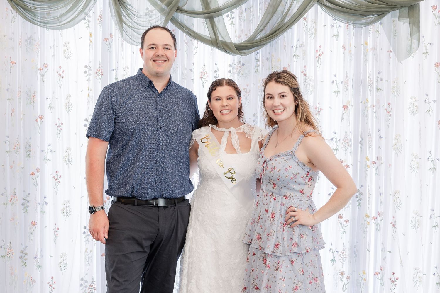 Bride in white wedding dress posing with two guests in front of elegant draped curtain backdrop.