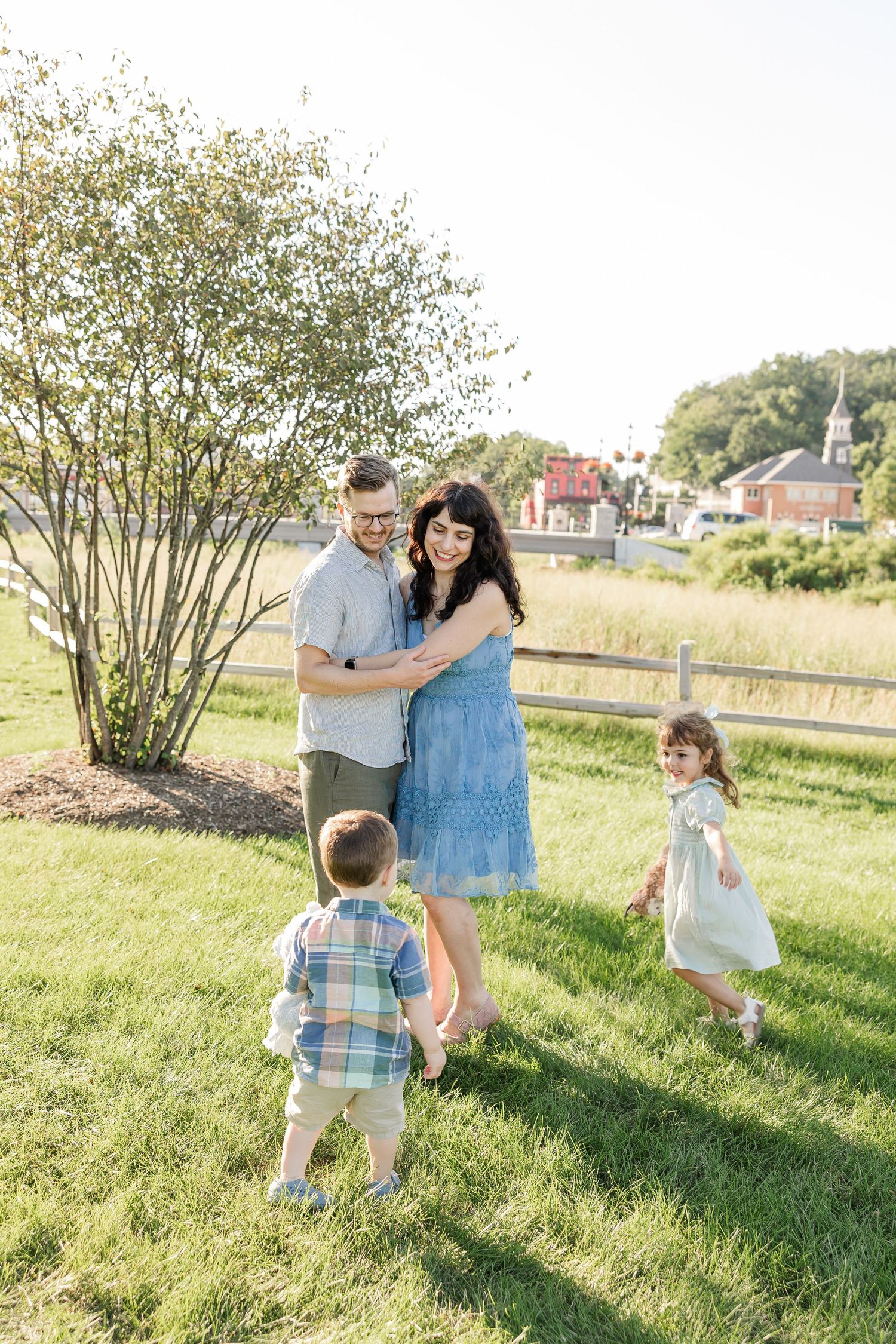 A happy family with two young children enjoy a sunny outdoor session near a wooden fence and blooming shrub.