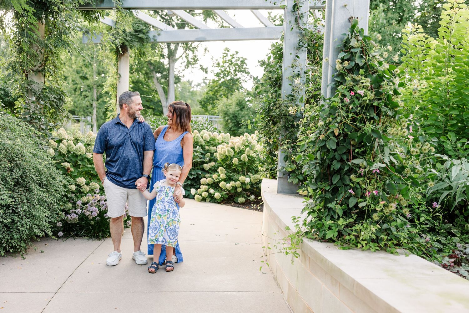 Family of three walking together under a pergola surrounded by lush green garden foliage.