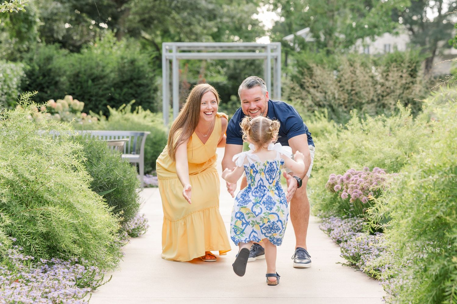 Happy family with toddler running toward parents on sunny garden path with lush green plants.