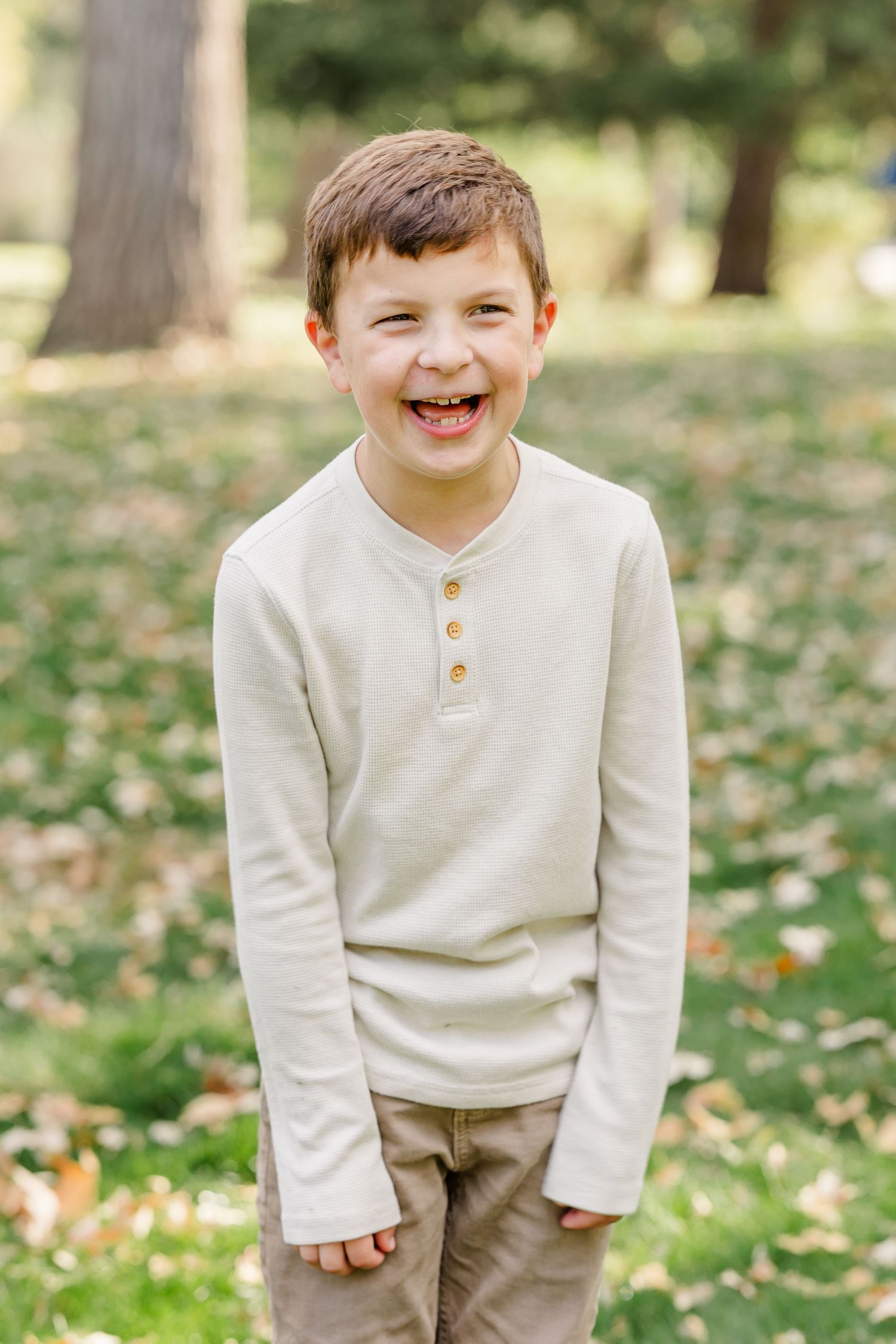 A happy young boy laughing outdoors in a park, wearing a cream henley shirt and khaki pants among green grass.