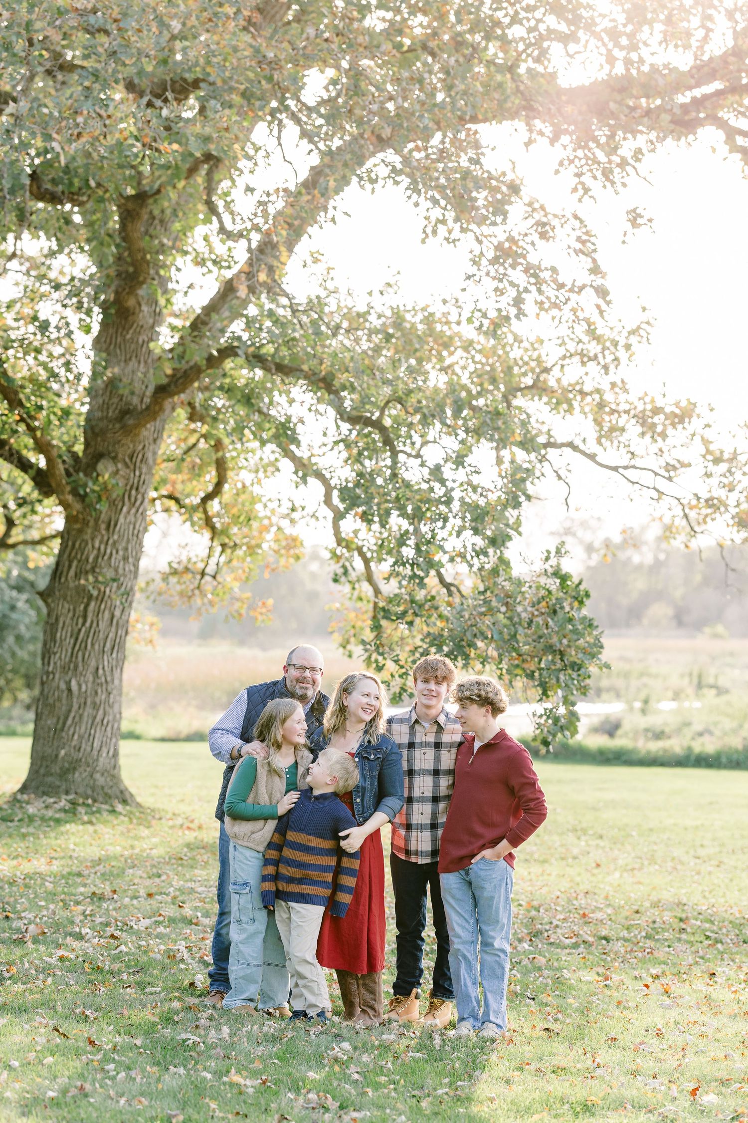 Family of six laughing together beneath a large oak tree on a grassy lawn during a warm autumn outdoor portrait session.