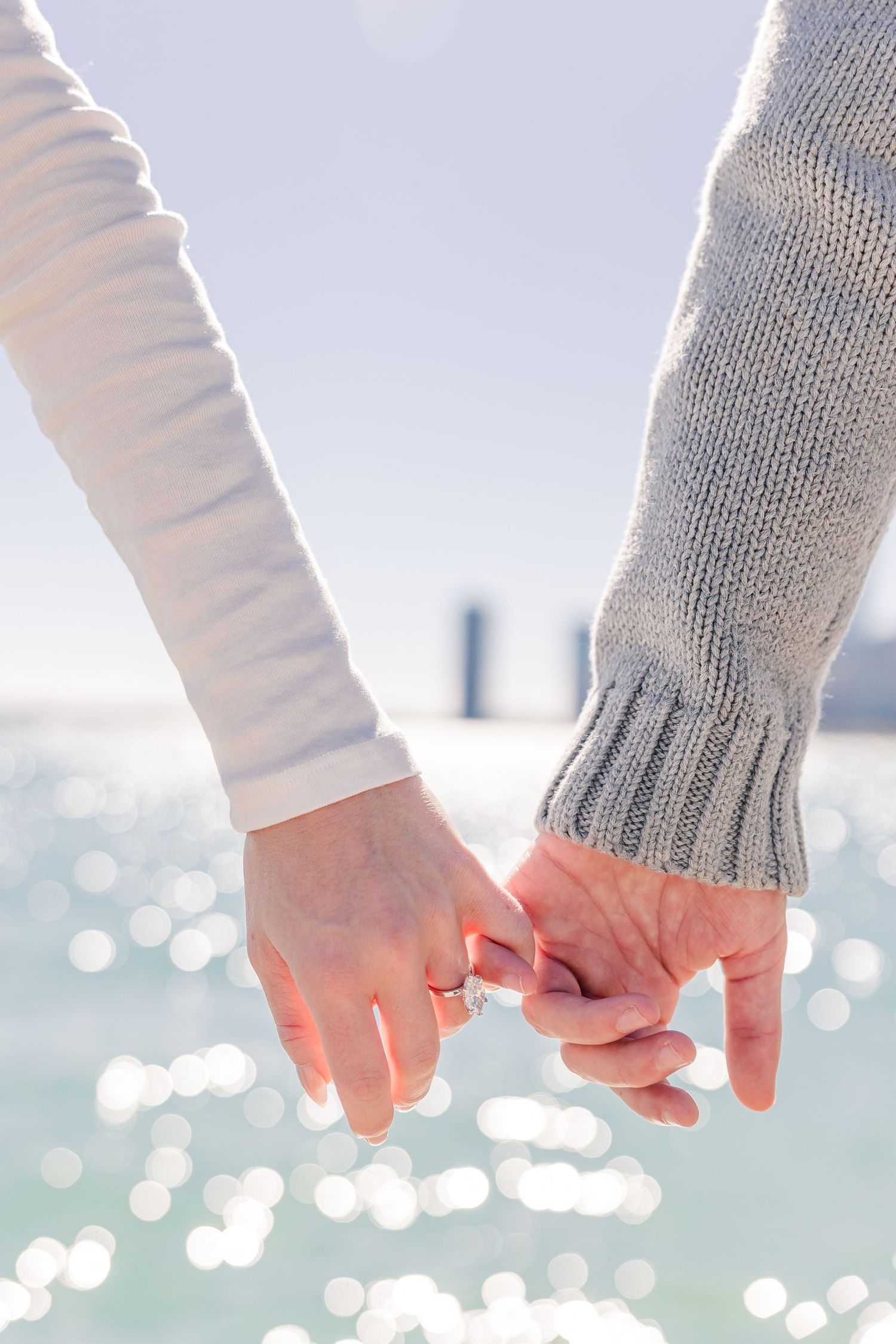 Close-up of couple holding pinky fingers together with sparkling water and city skyline softly blurred behind them.