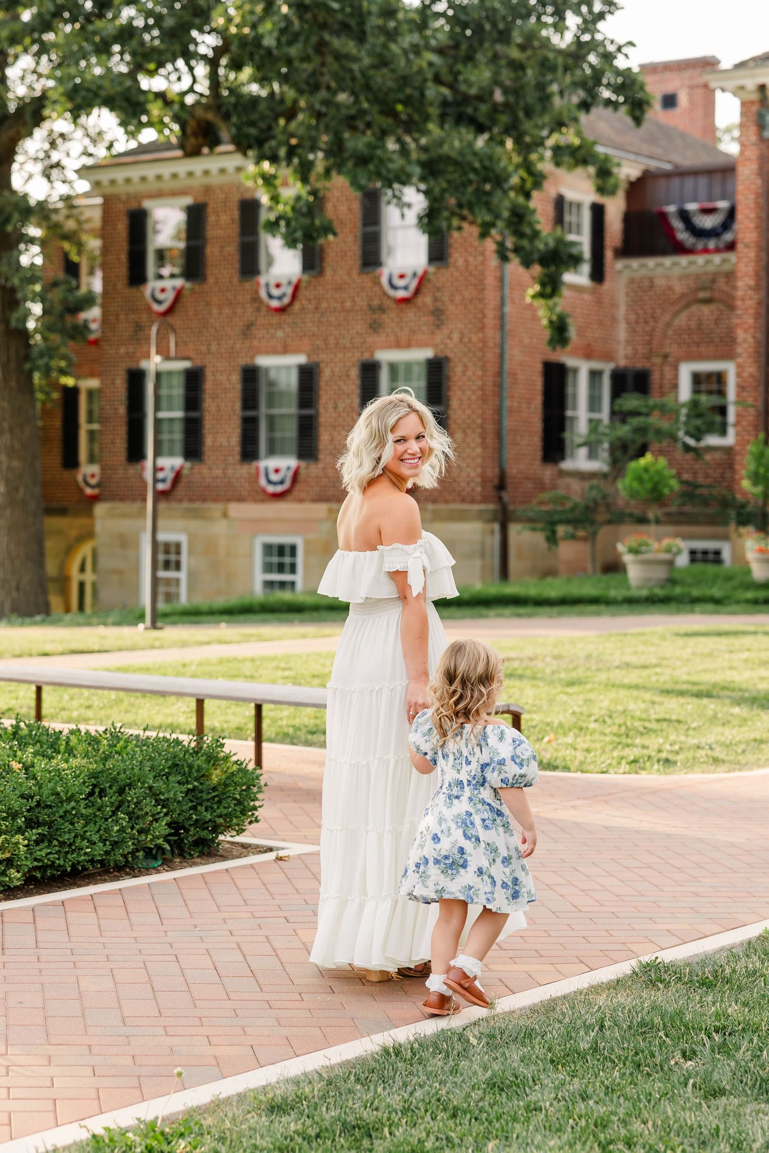 A smiling blonde woman in a white off-shoulder dress holds hands with a little girl in a floral dress on a brick path.