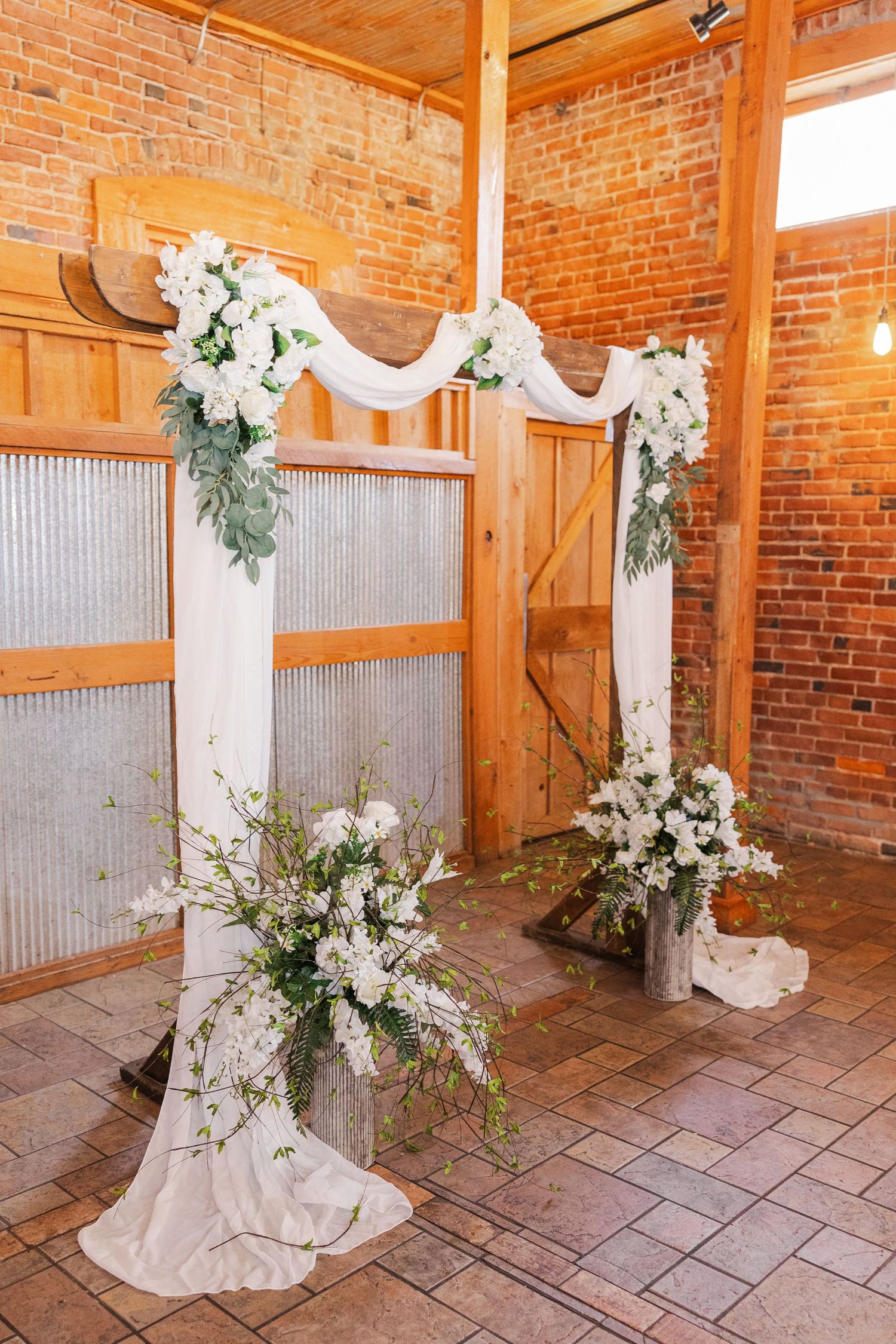White wedding arch with flowing fabric and floral arrangements against exposed brick wall.