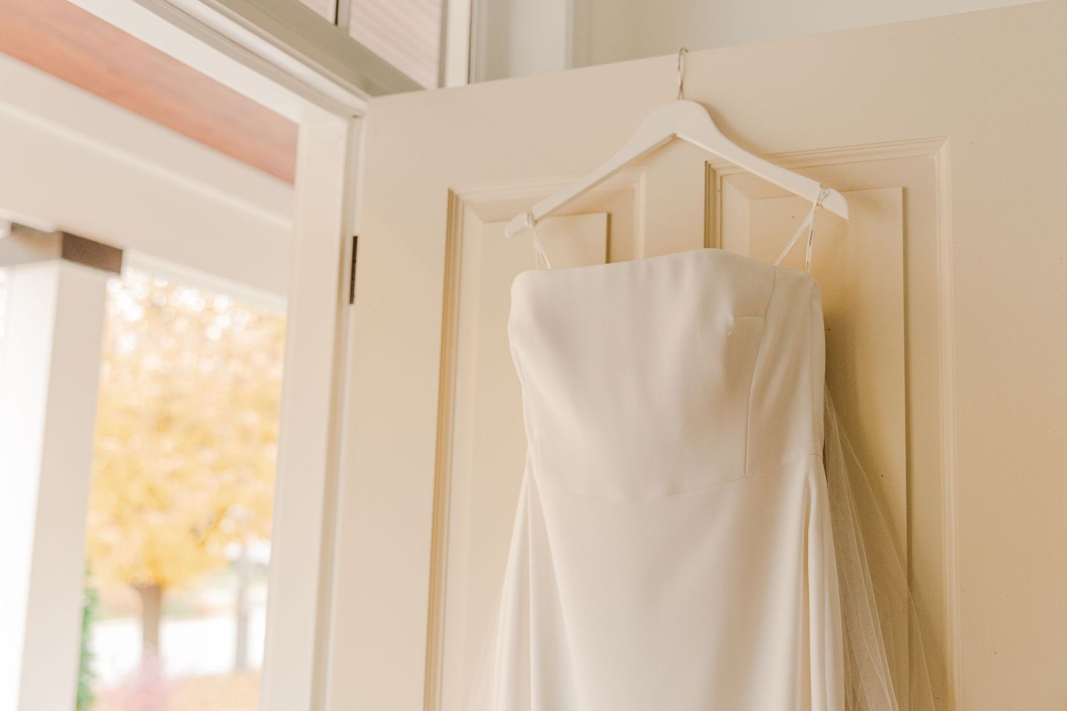 White bridal gown with clean lines displayed on a hanger against an ivory wall.