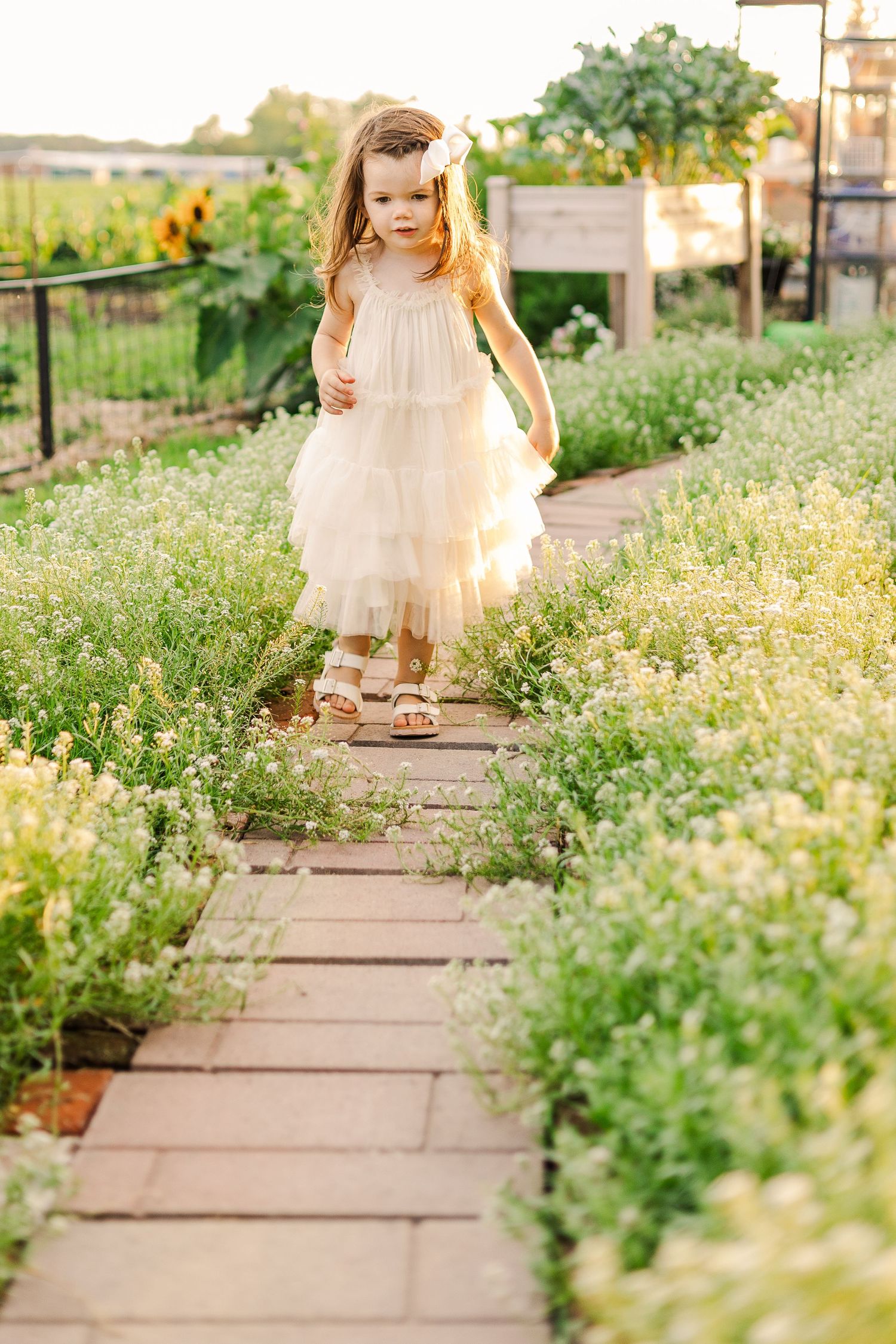 A delicate white dress twirls down a garden pathway lined with wildflowers at sunset.
