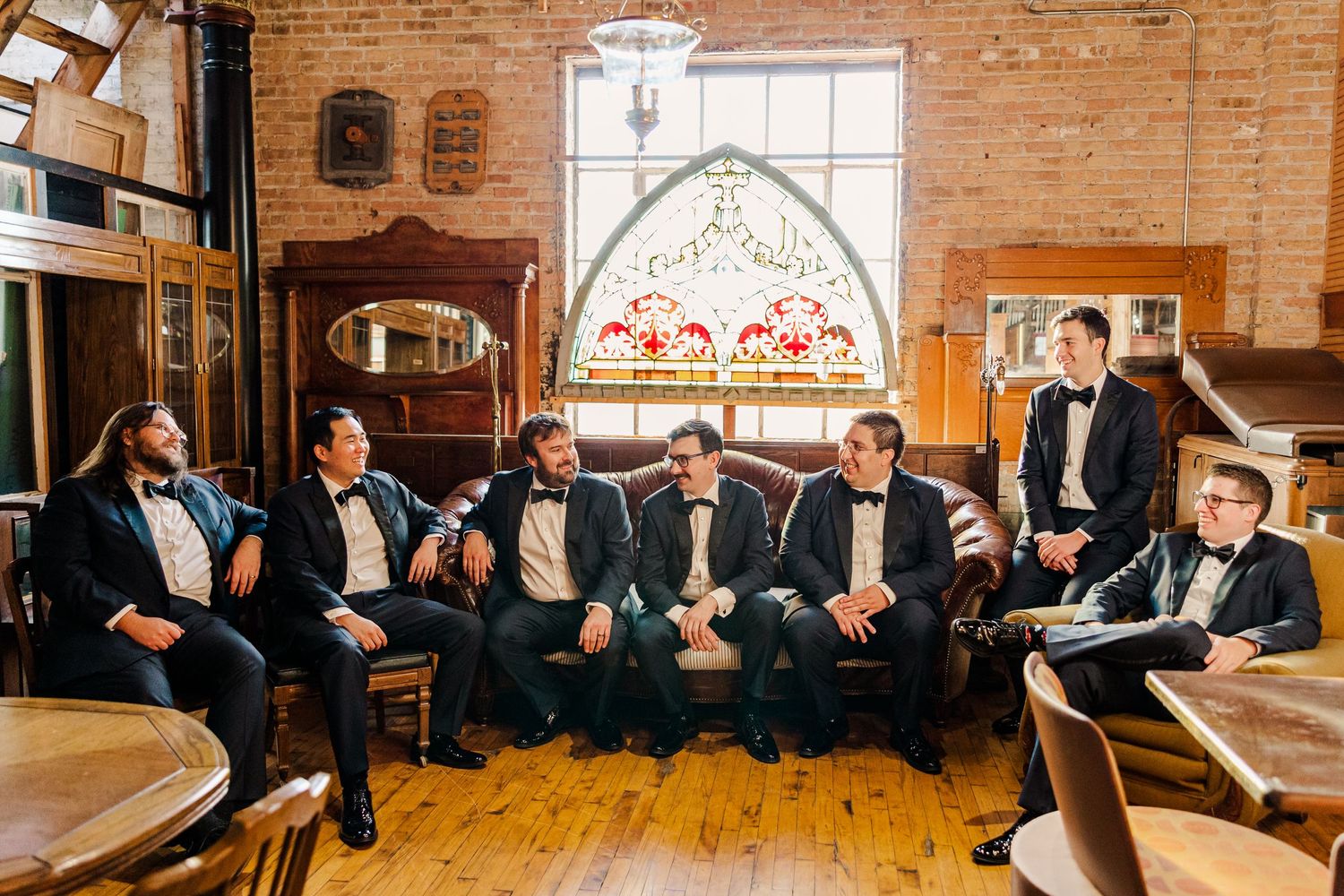 Groomsmen in formal attire sit together on vintage furniture in a rustic brick-walled room with arched stained glass window.