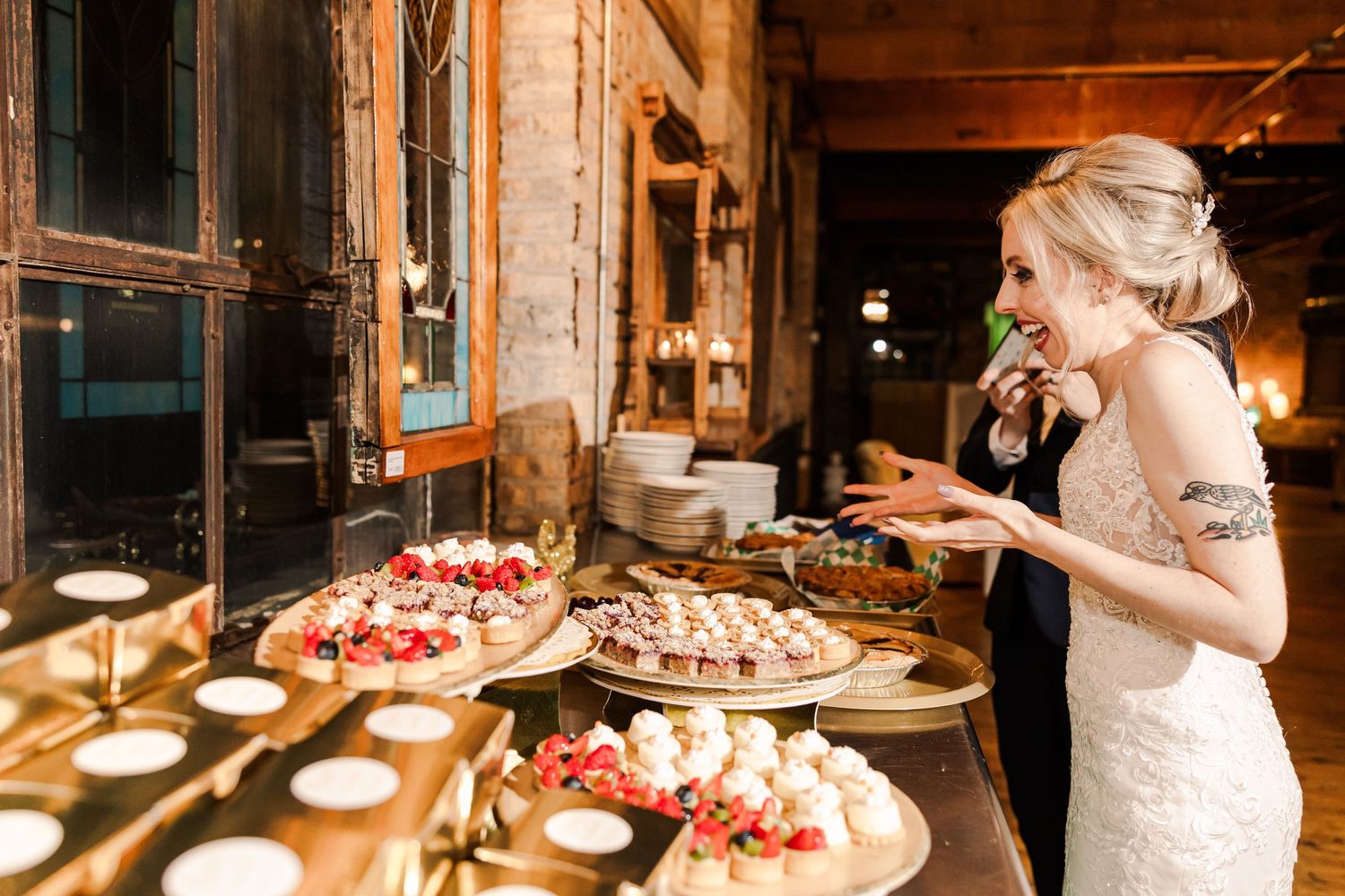 Wedding reception dessert table features assorted cakes and treats with rustic brick wall backdrop.