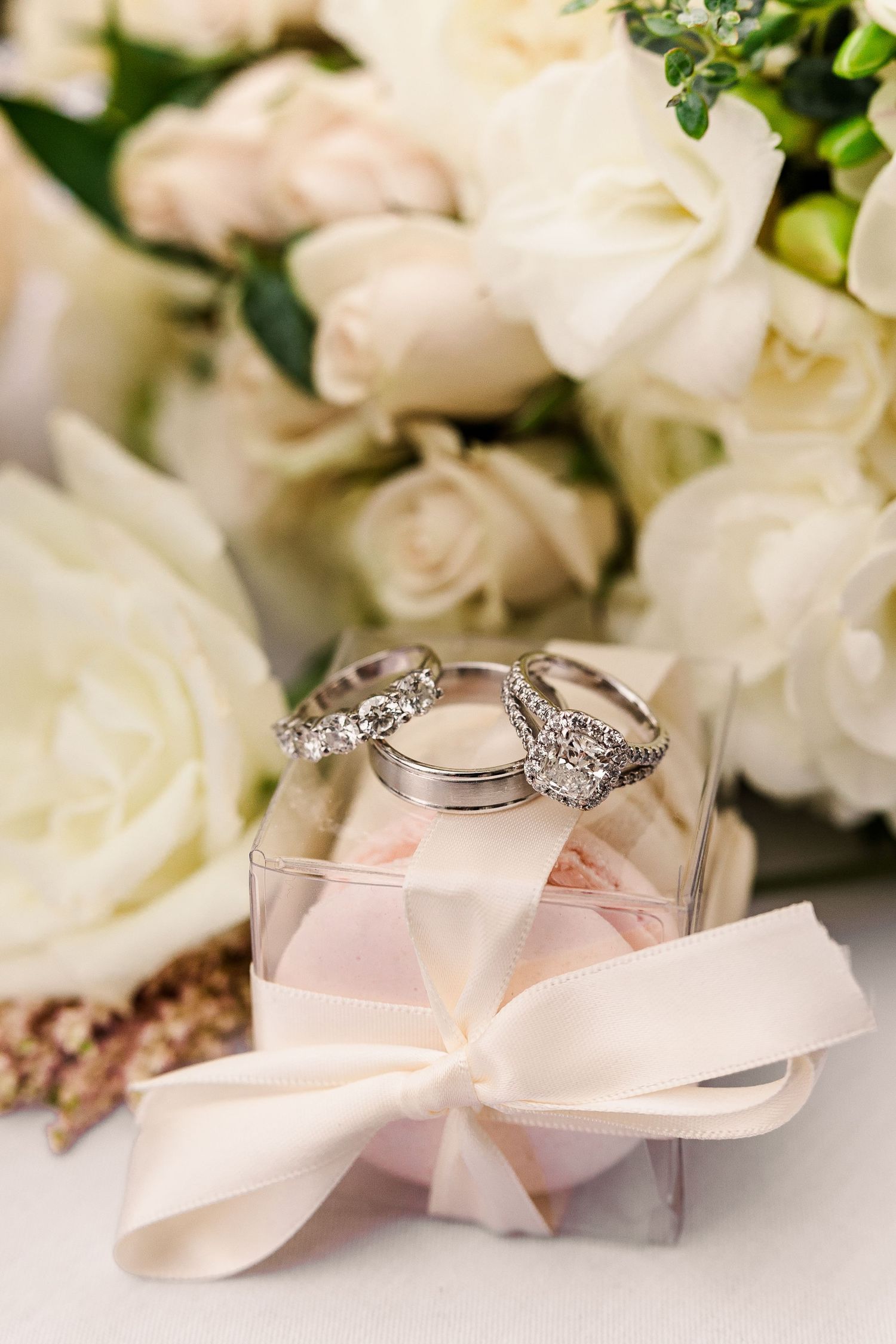 Wedding rings displayed on decorative box with blush bow and white rose bouquet in background.