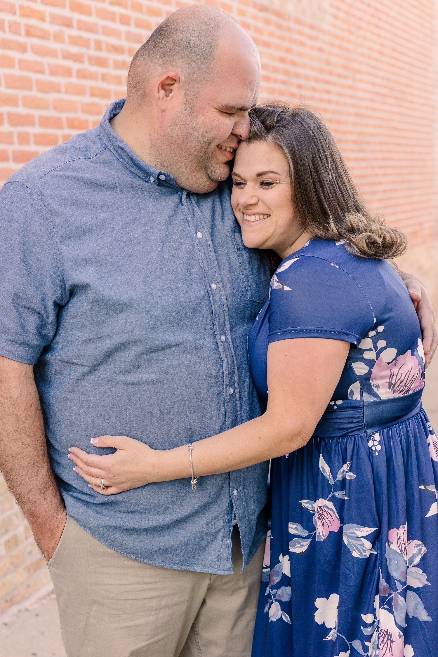 Couple embracing against brick wall wearing blue shirt and floral dress.