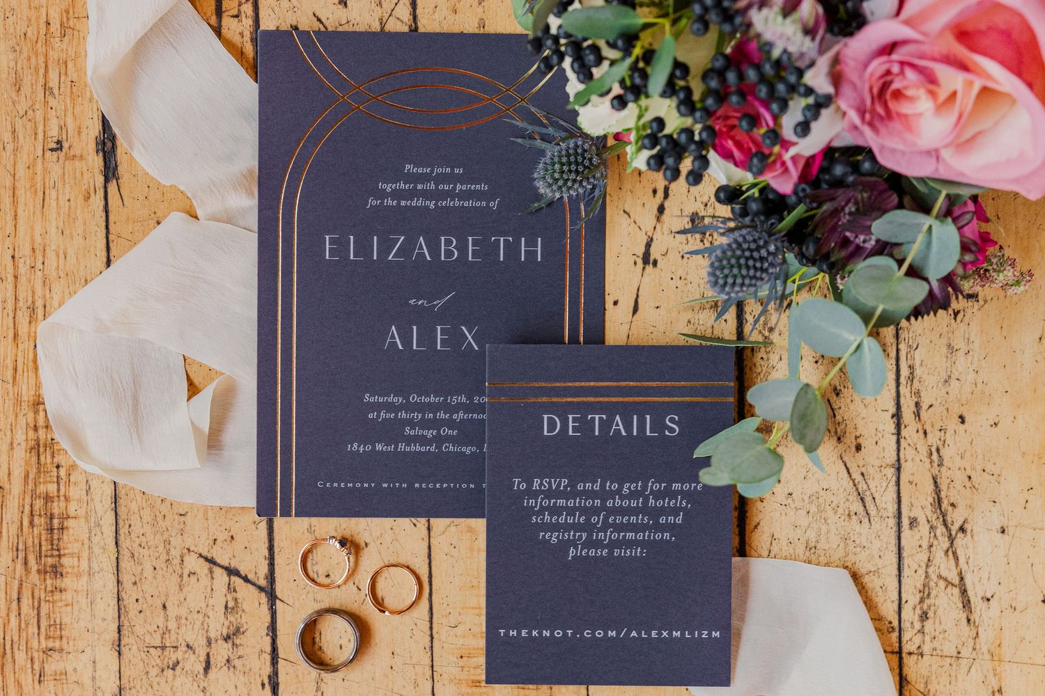 Wedding stationery suite in navy blue with metallic details alongside wedding rings and pink florals.