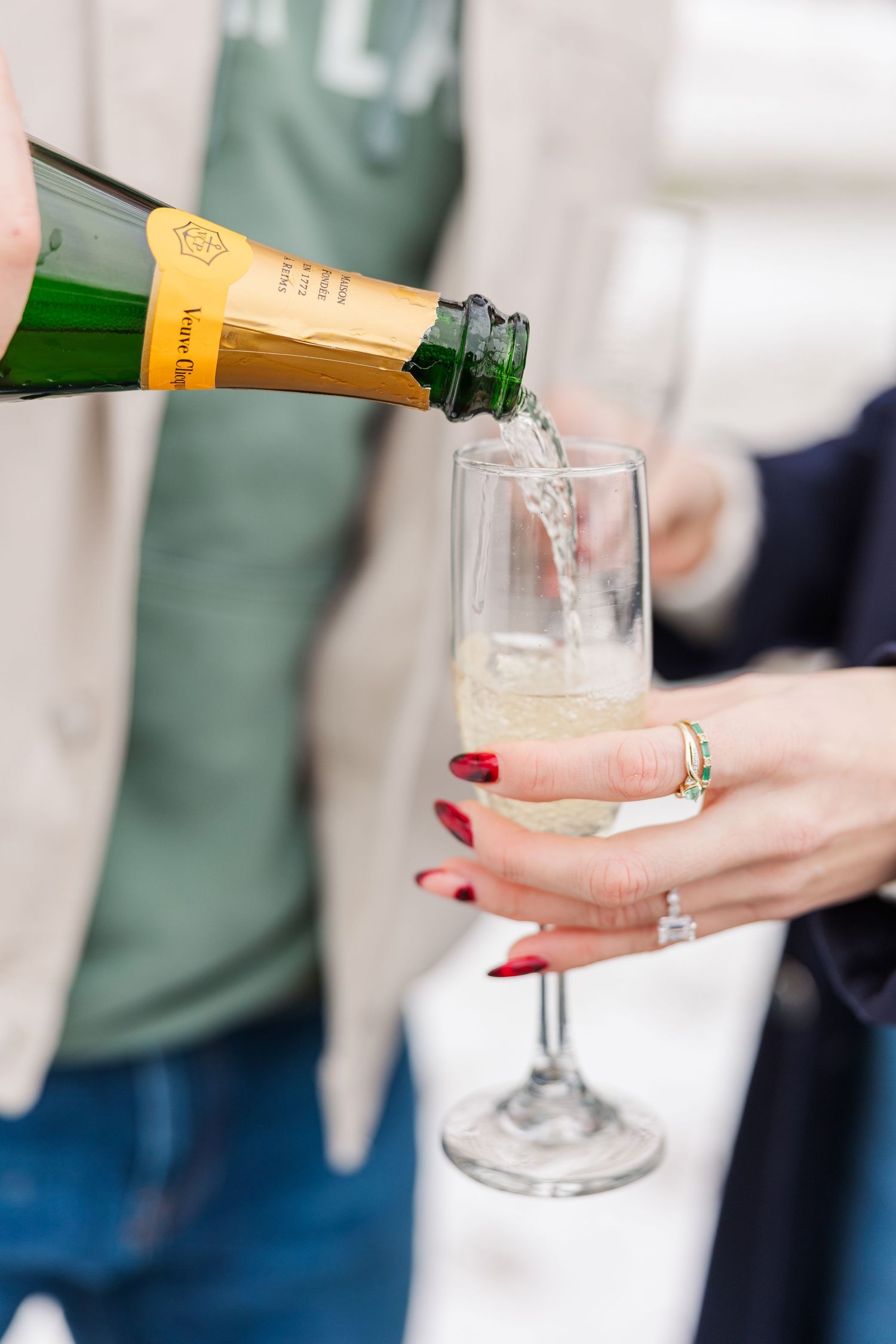 Champagne being poured into a glass flute held by a hand with red nail polish and gold rings.