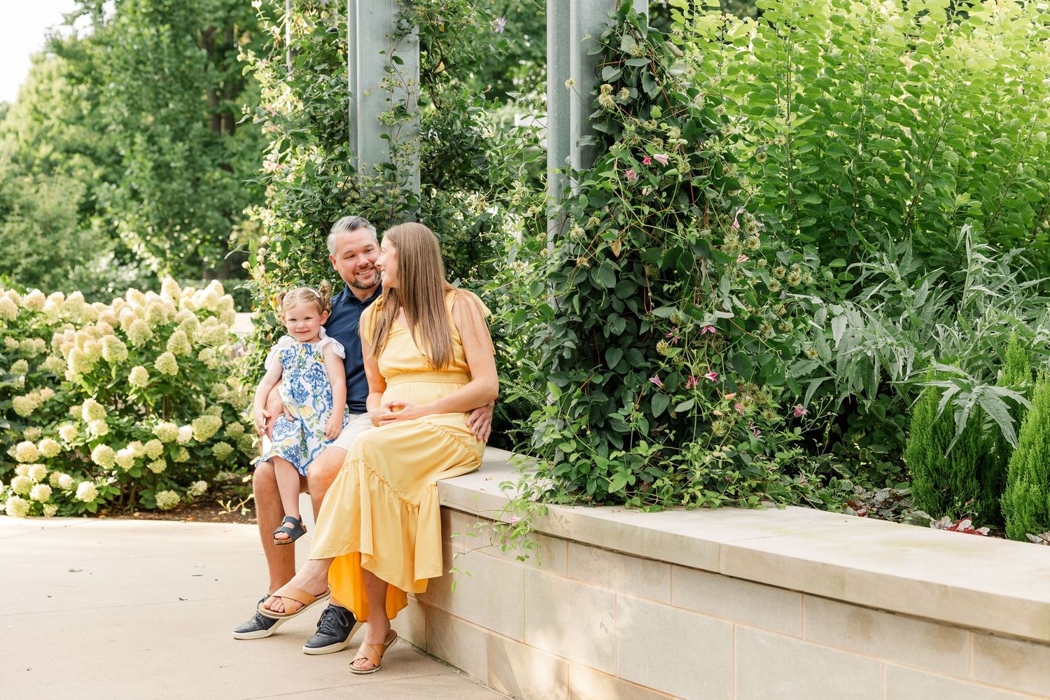 Pregnant woman in yellow dress sits with partner and child on stone bench in lush garden for maternity photo.