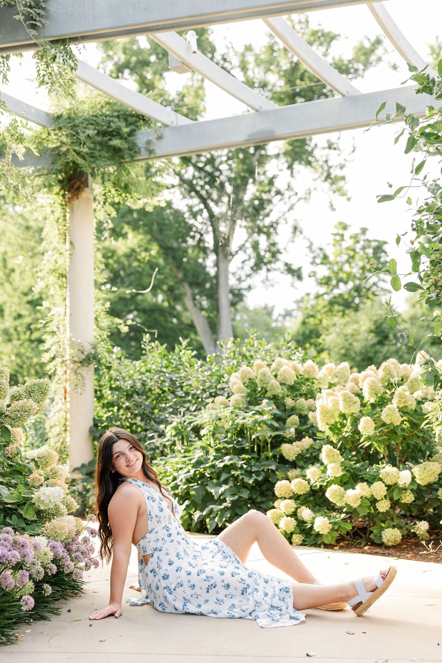 Young woman in a blue floral dress sitting under a pergola surrounded by blooming hydrangeas in a garden.