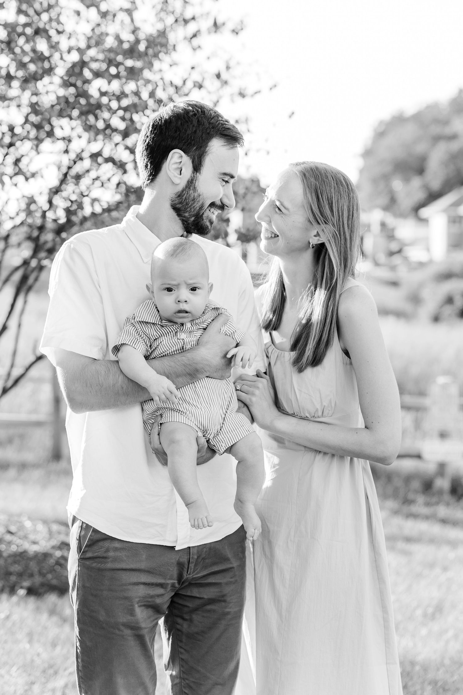 Black and white photo of a young couple smiling at each other while man holds their baby outdoors.