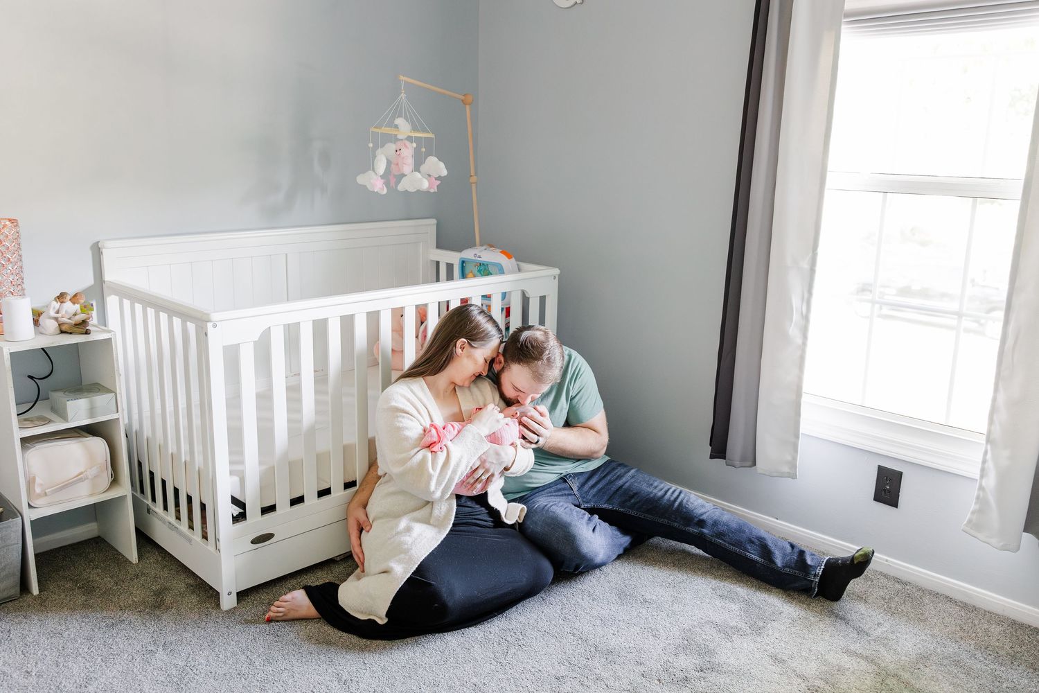 Young parents sitting on nursery floor with newborn baby next to white crib.