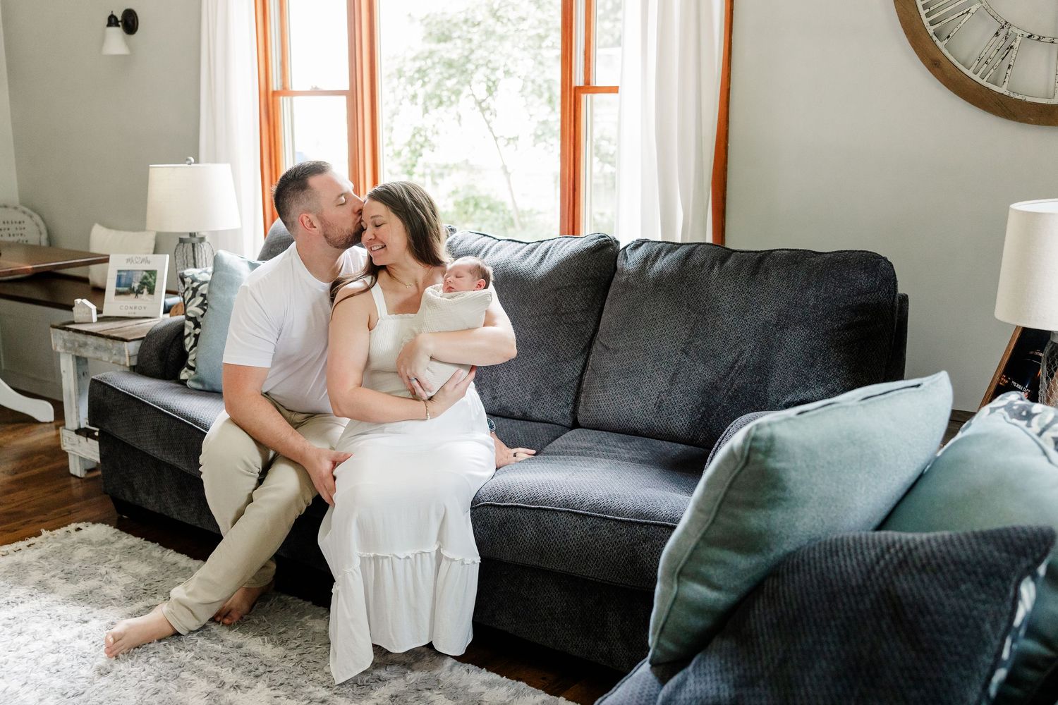 A couple sits on a gray sofa, sharing a kiss while cradling their newborn baby in a cozy living room.