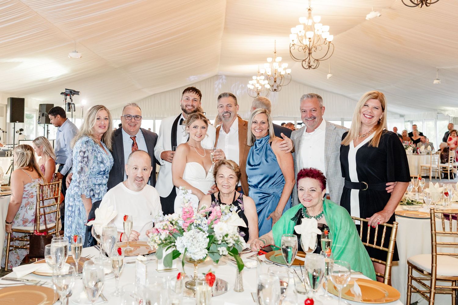 Wedding party and family members pose together smiling at a floral-decorated reception dinner table.