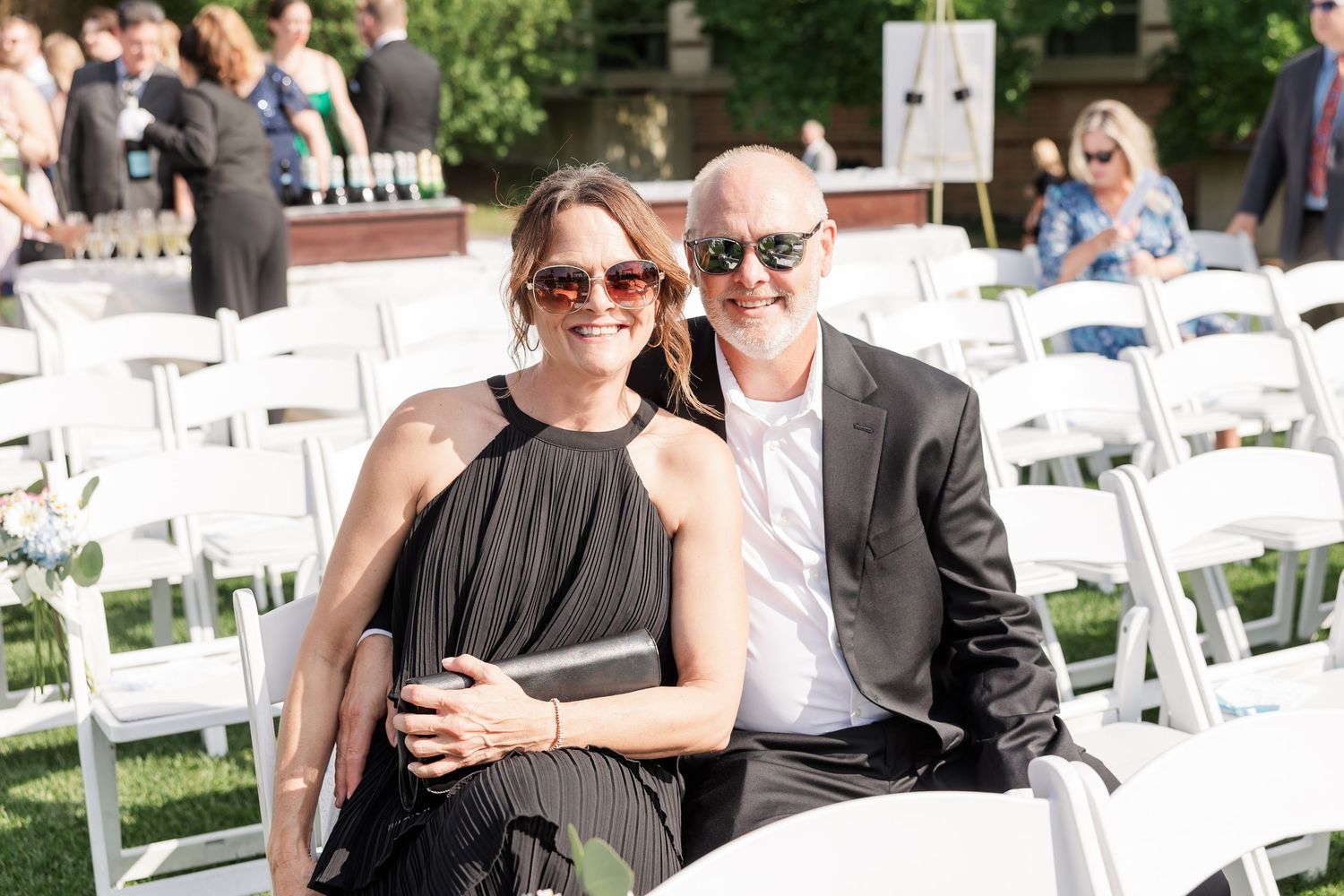 Smiling couple in formal attire seated in white chairs at an outdoor wedding ceremony.