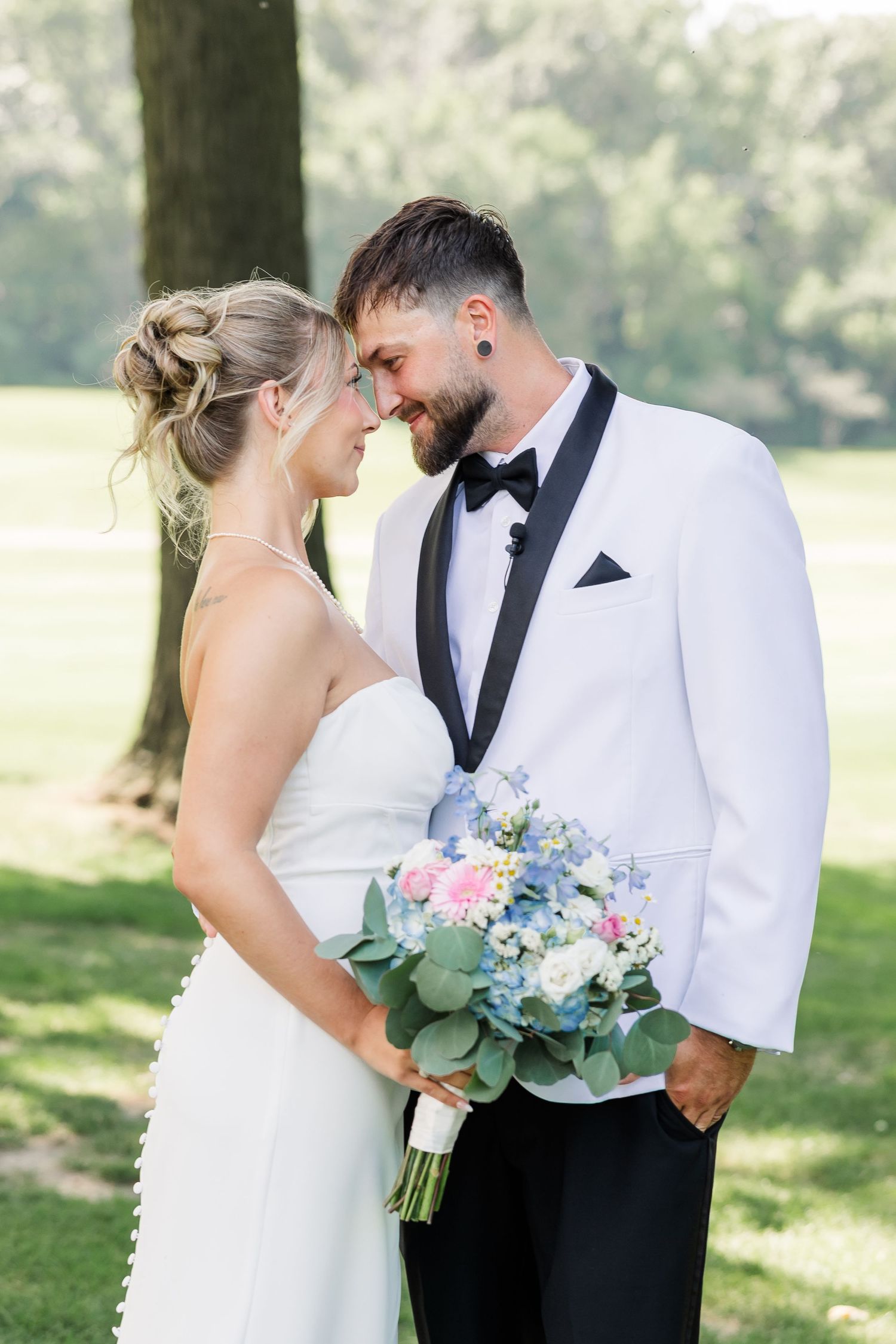 Bride and groom in white tuxedo share romantic forehead touch outdoors holding blue and pink wedding bouquet.