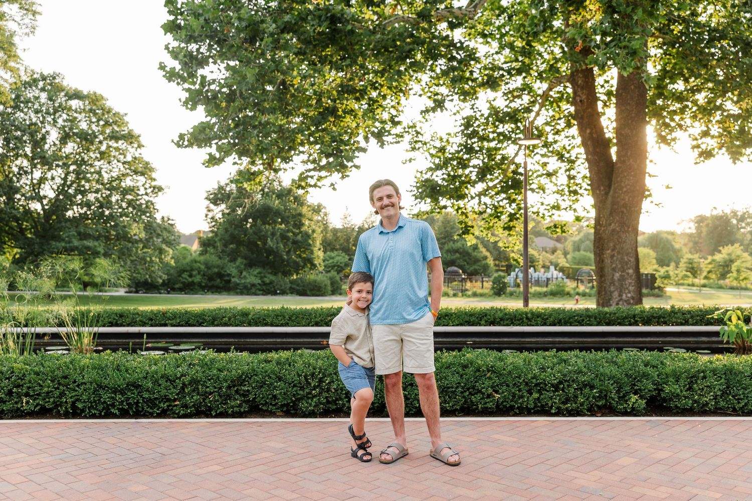 Father and young son smiling together on a brick path surrounded by green hedges and trees.