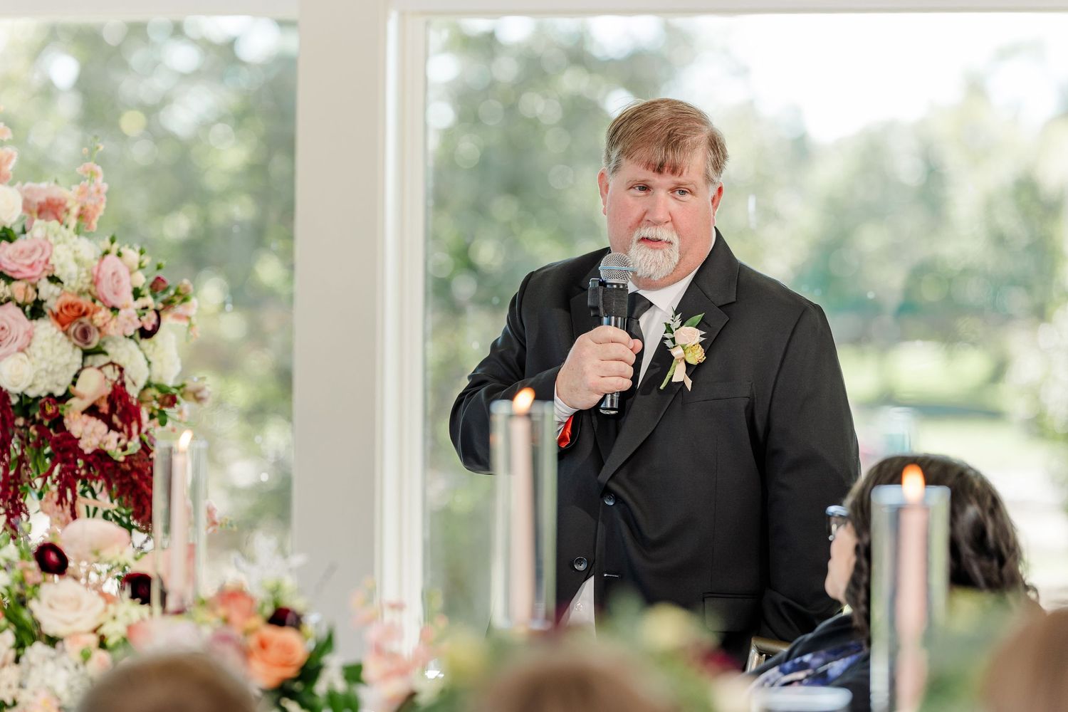 Man in a dark suit with a boutonniere gives a wedding toast into a microphone beside candles and colorful floral centerpieces.