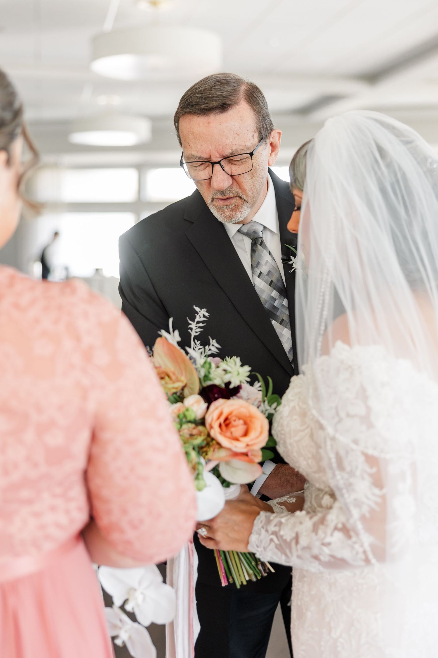 Bride in a lace gown and veil holds a peach rose bouquet while a man in glasses and a dark suit leans in during a wedding ceremony.