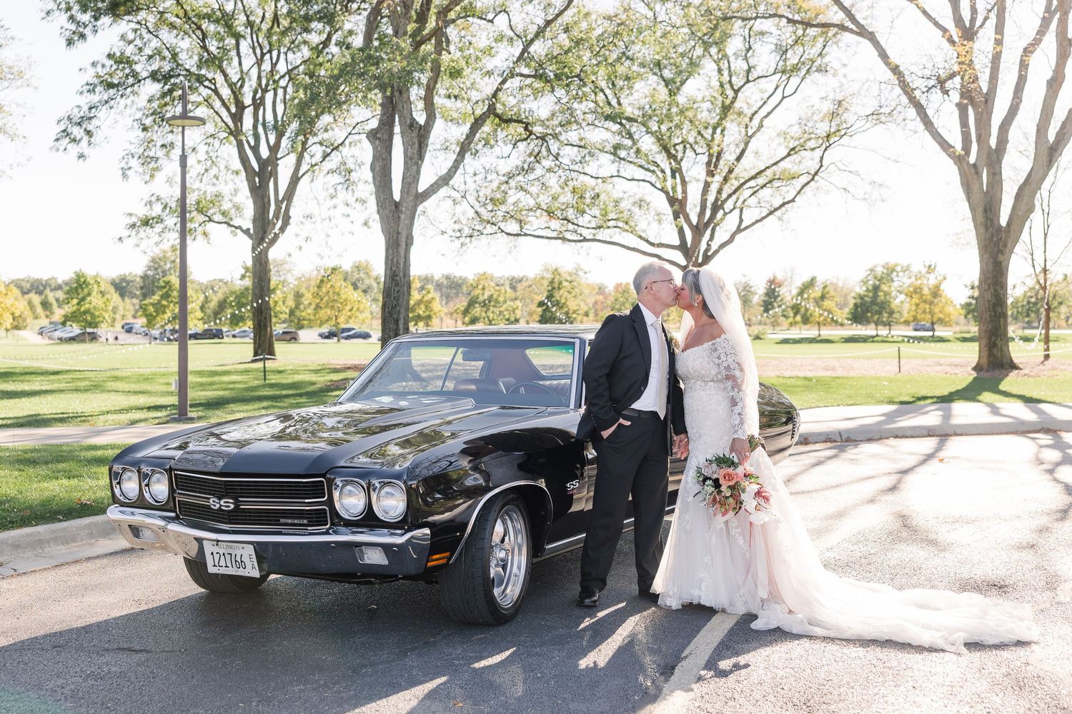 Bride in a white gown and groom in a dark suit embrace beside a classic black muscle car on a sunny tree-lined road.
