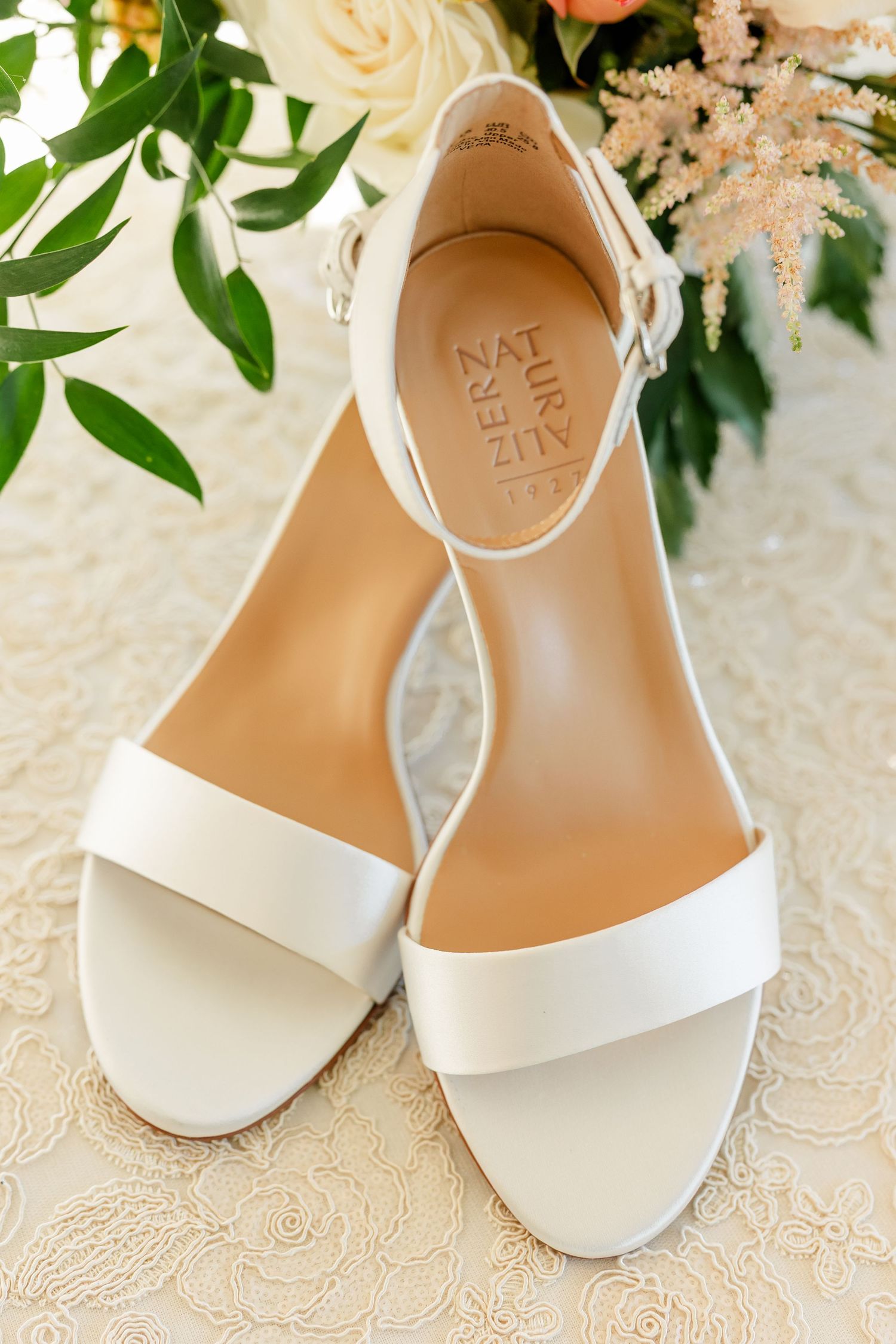 Ivory Naturalizer ankle-strap block heel sandals resting on delicate lace beside a white rose and green foliage bridal bouquet.