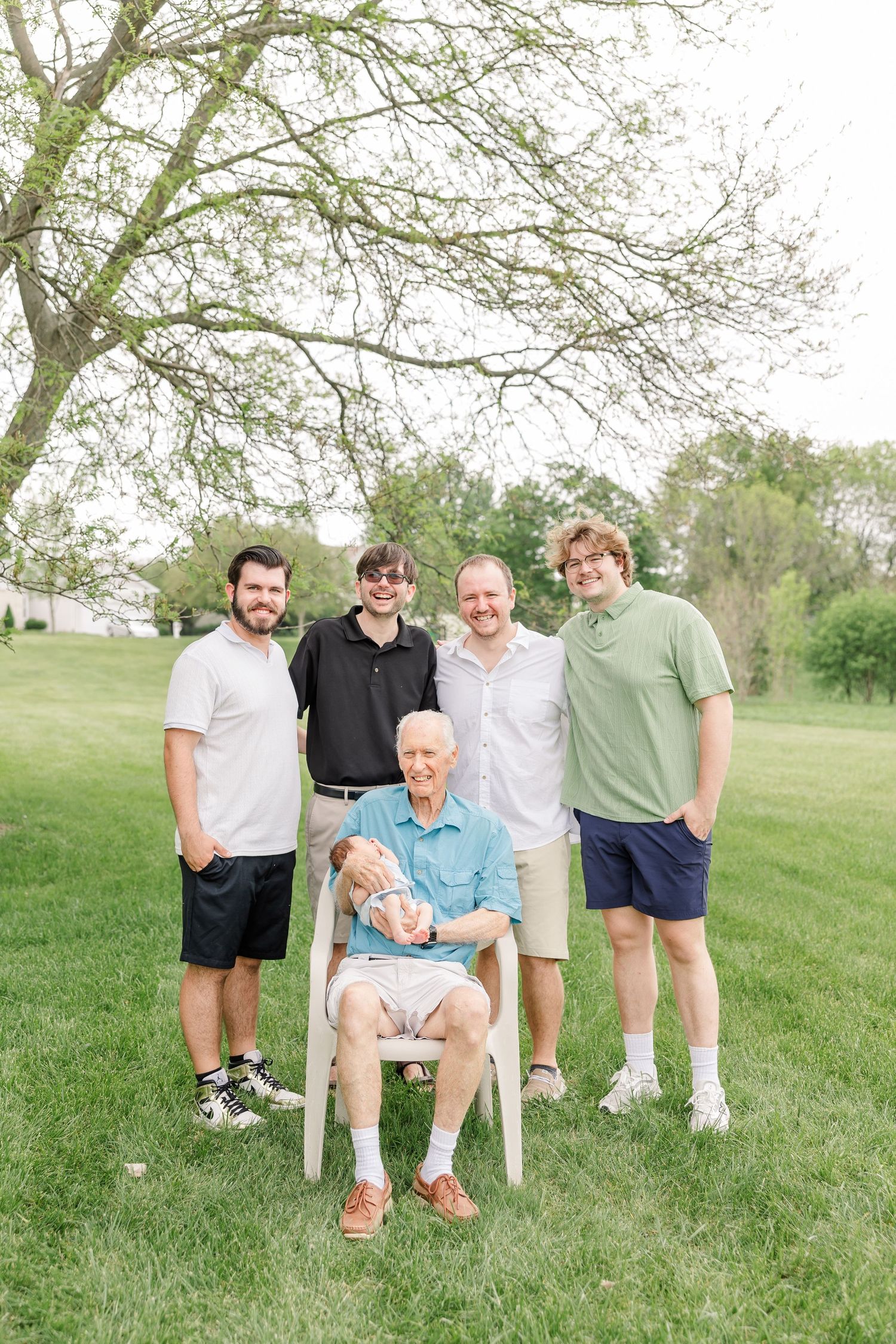 Elderly grandfather seated holding newborn surrounded by four adult grandsons in a park setting.