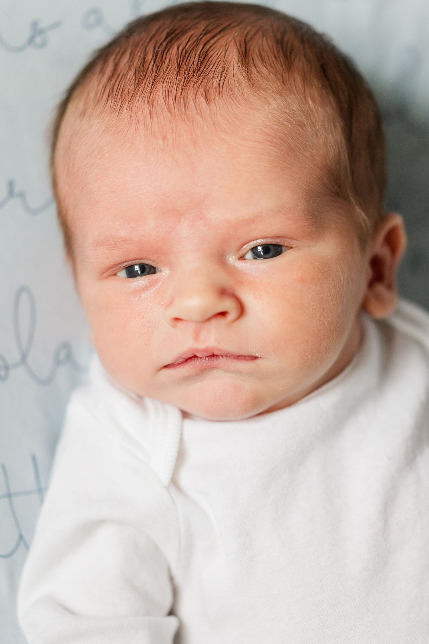 Close-up portrait of alert newborn baby with dark eyes wearing white onesie on blue blanket.