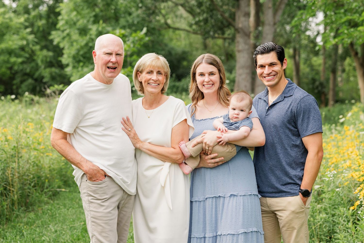 Multi-generational family of five smiling outdoors in a green park, with grandparents, parents, and a baby.