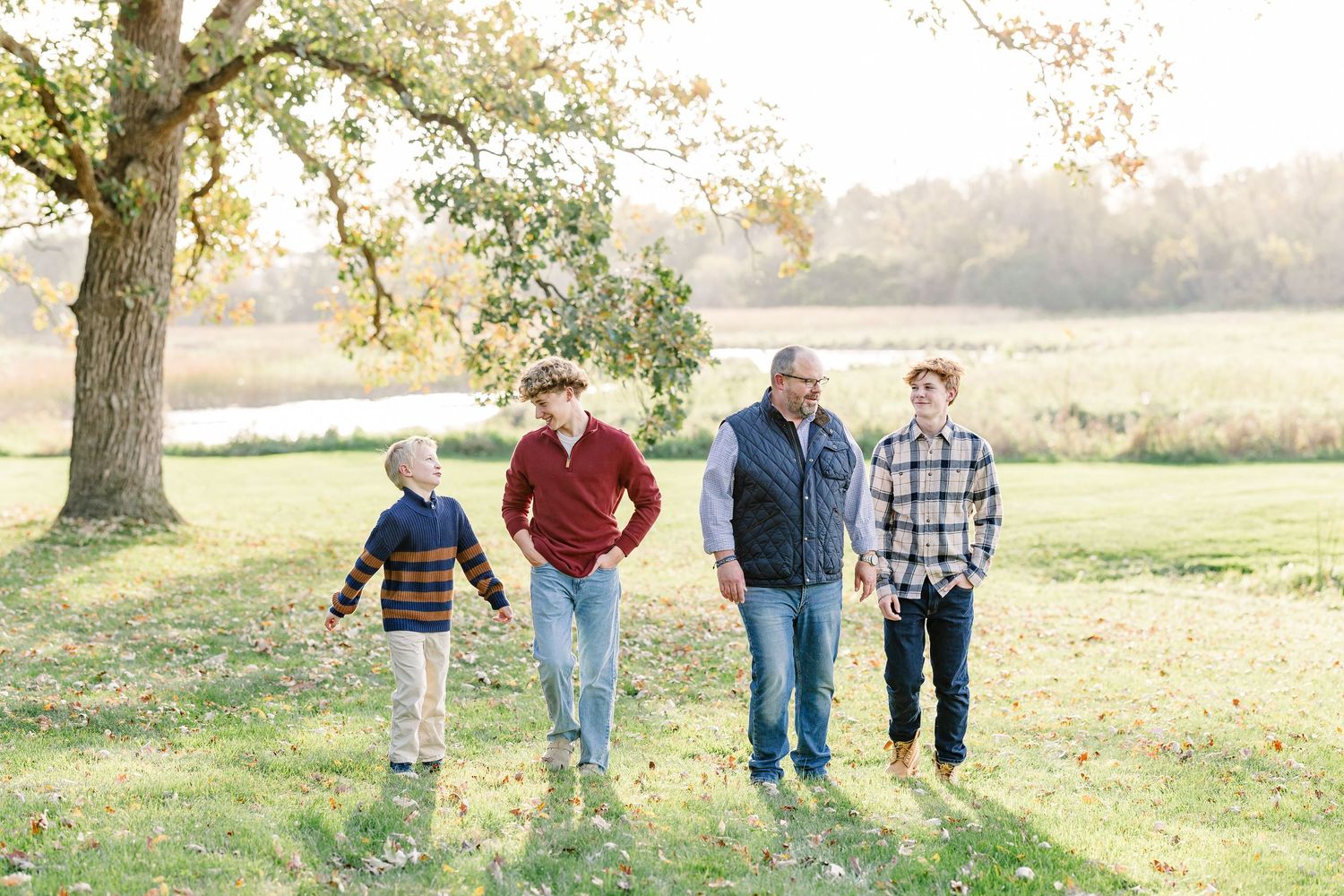 Three adults and a young boy walk together across a grassy park beneath a large tree during a golden autumn family photo session.