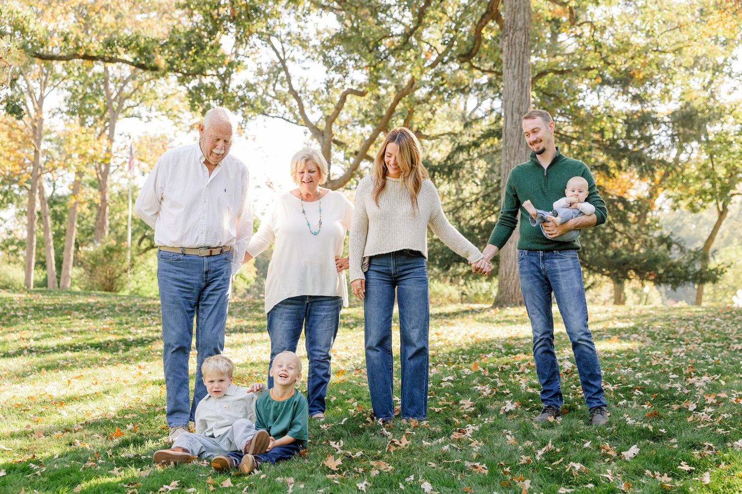Multi-generational family of six poses outdoors in autumn park, holding hands with children, wearing jeans and casual tops.