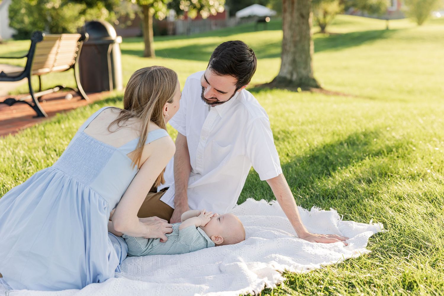 A young couple sits on a white blanket in a sunny park, smiling at their baby lying between them.