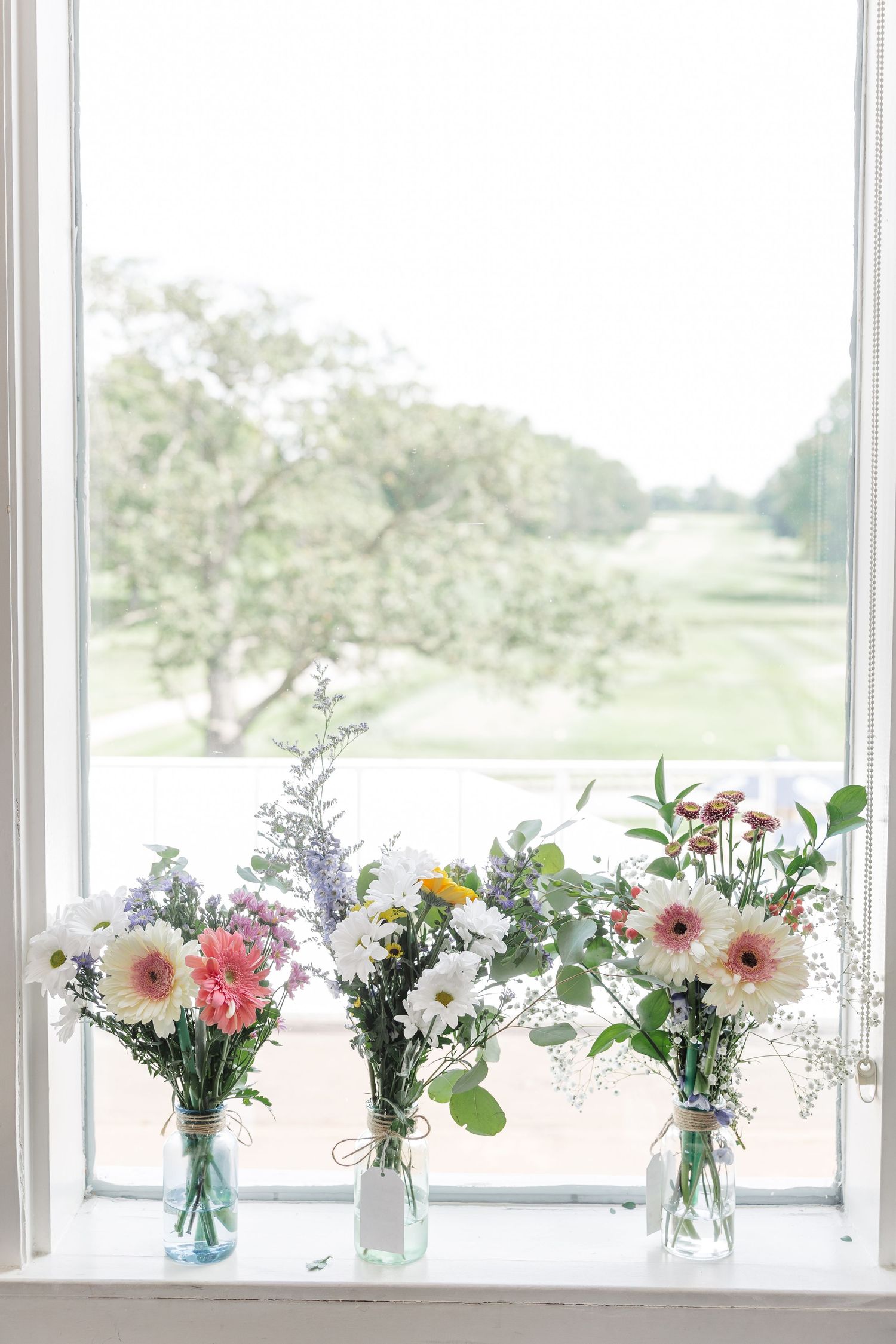 Three small bouquets of gerbera daisies and wildflowers in glass jars on a bright window sill.