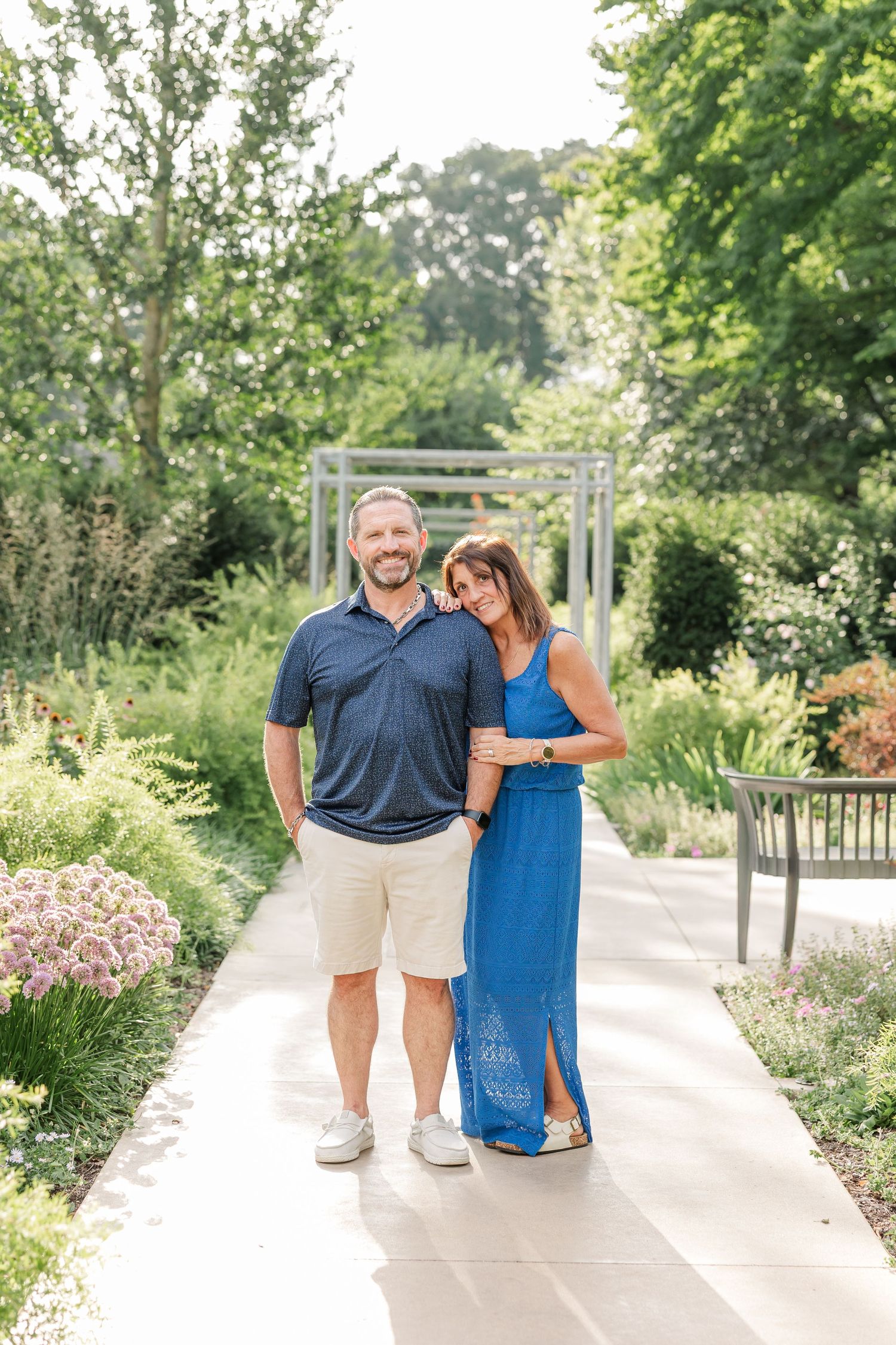 Couple smiling together on a garden path, man in navy polo and khaki shorts, woman in blue maxi dress.