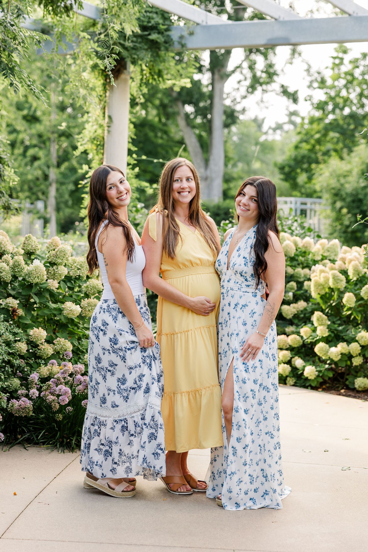 Three women pose in a garden, the center one pregnant in yellow dress, flanked by two in blue floral maxi dresses.