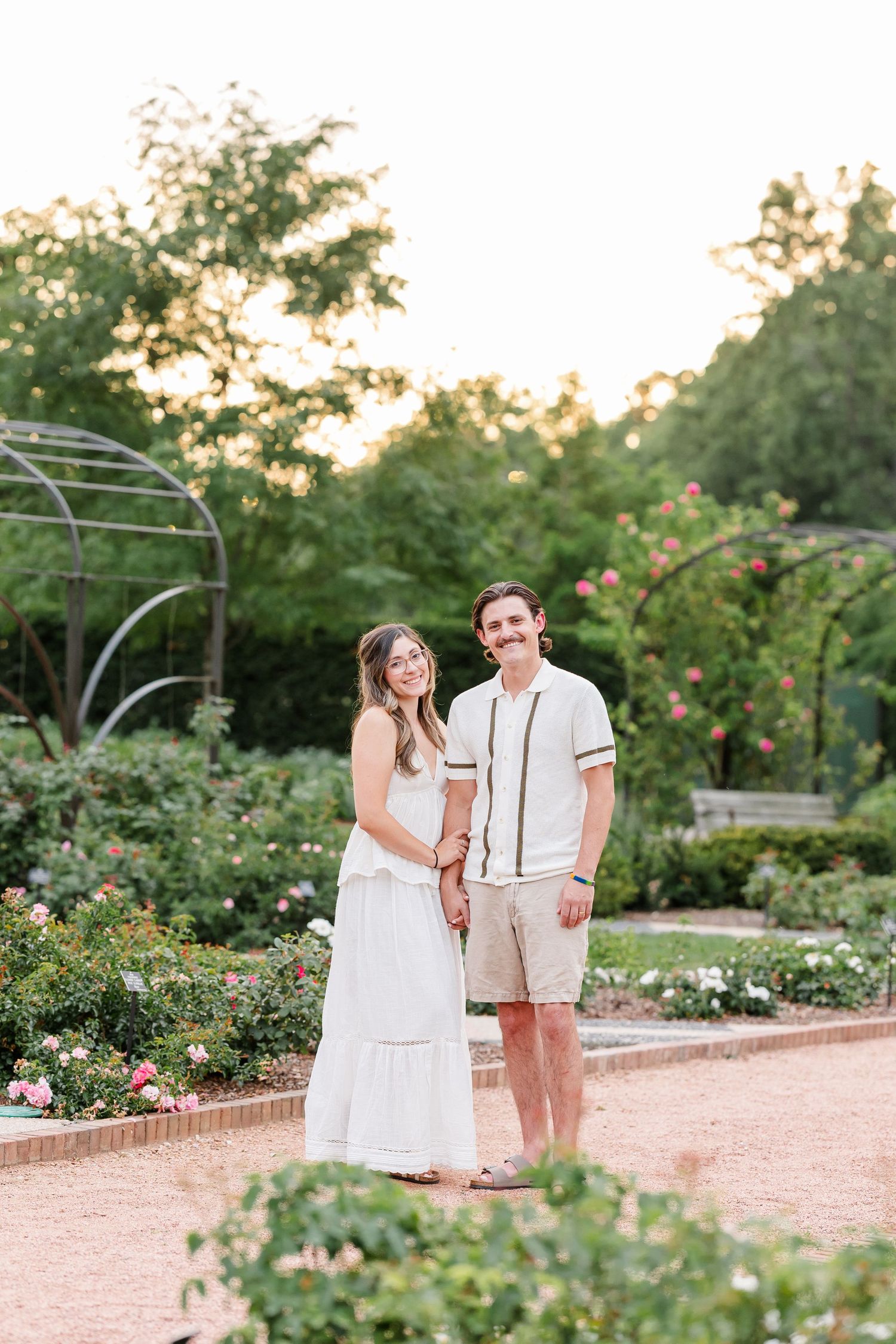 Couple in coordinating white outfits holds hands while posing in blooming rose garden at golden hour.