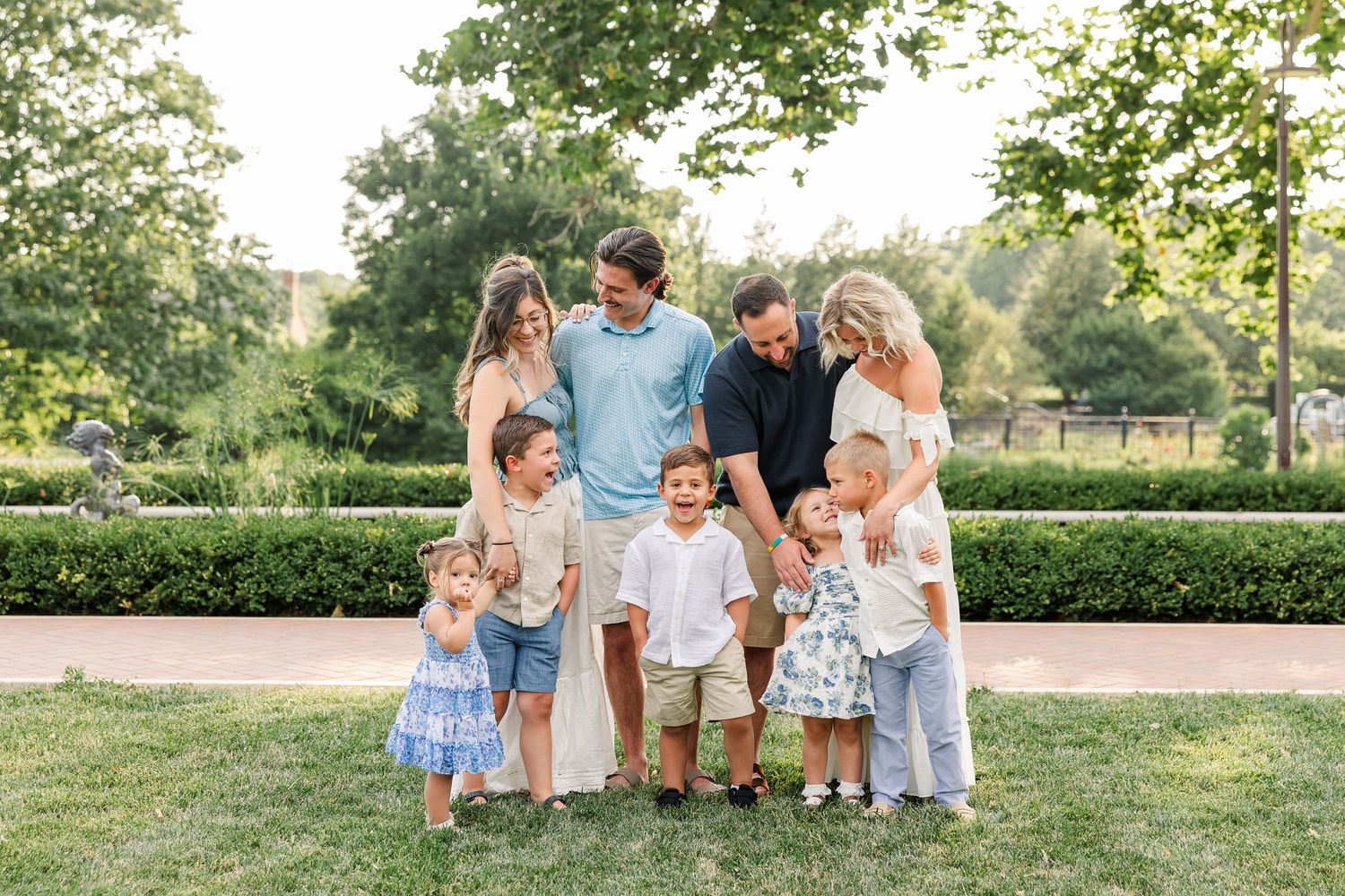 Large family group portrait outdoors in a lush green park, parents with children smiling together on a sunny day.
