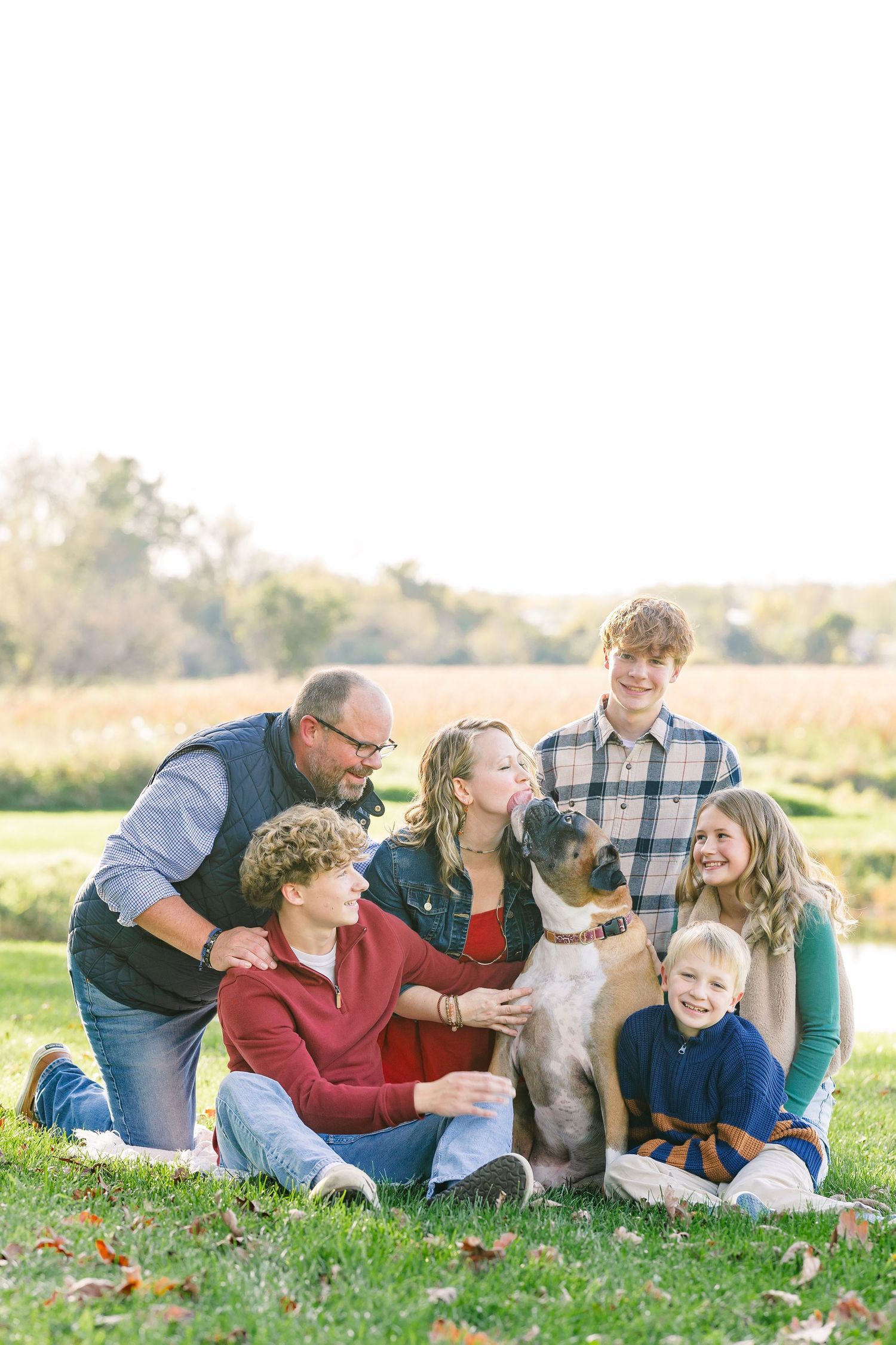 Family of six smiling and cuddling a large tan dog on the grass in a sunny field during an autumn outdoor portrait session.