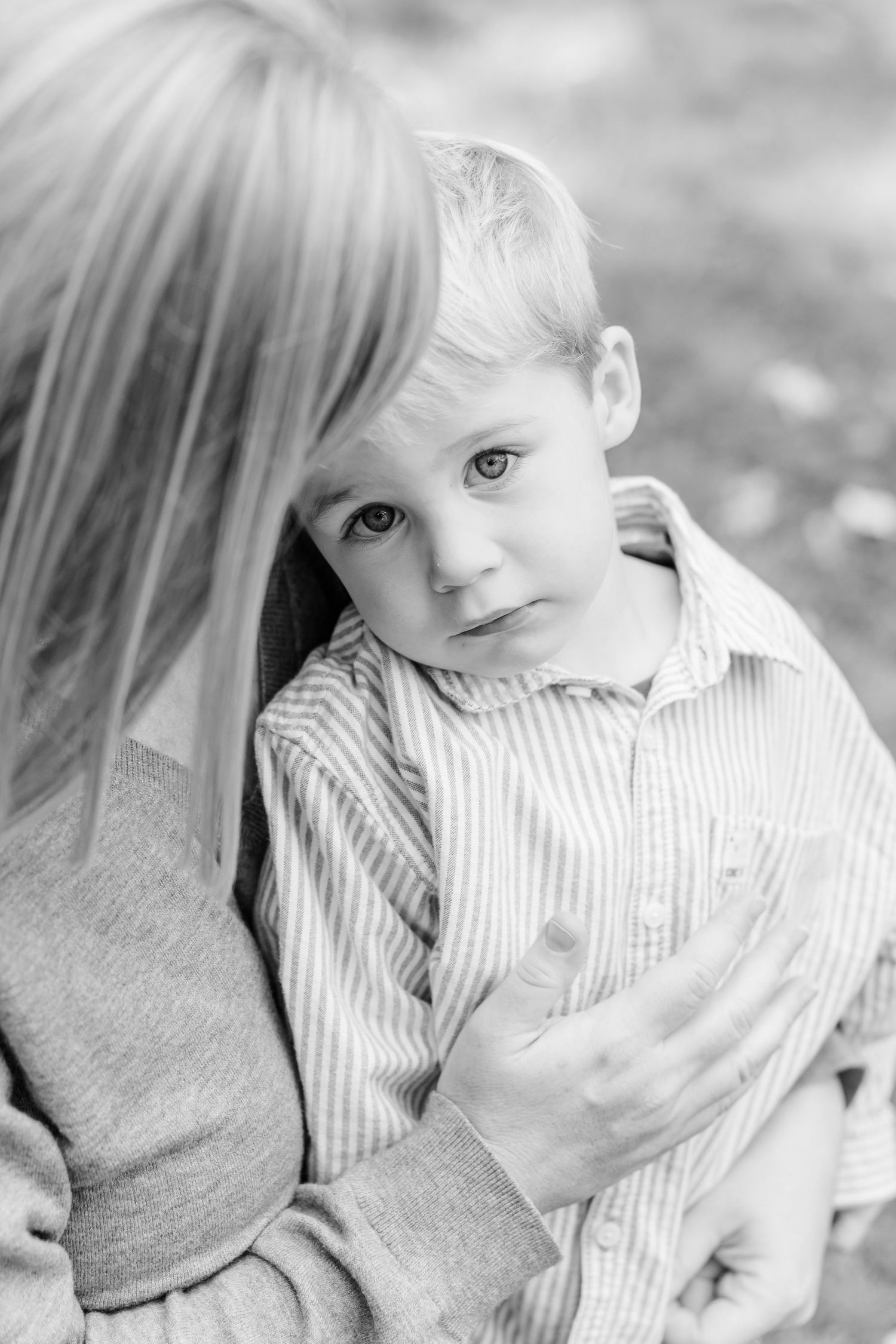 Black and white portrait of a young blonde boy being embraced by his mother outdoors.