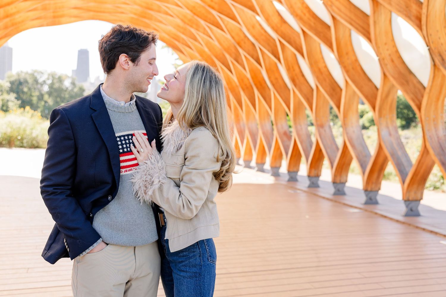 Couple sharing a romantic moment under a wooden arch structure in a park, man in navy blazer, woman in beige jacket.