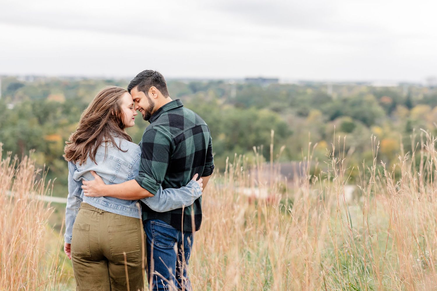 Couple embracing from behind in a tall golden grass field overlooking a wooded valley during an autumn engagement photo session.