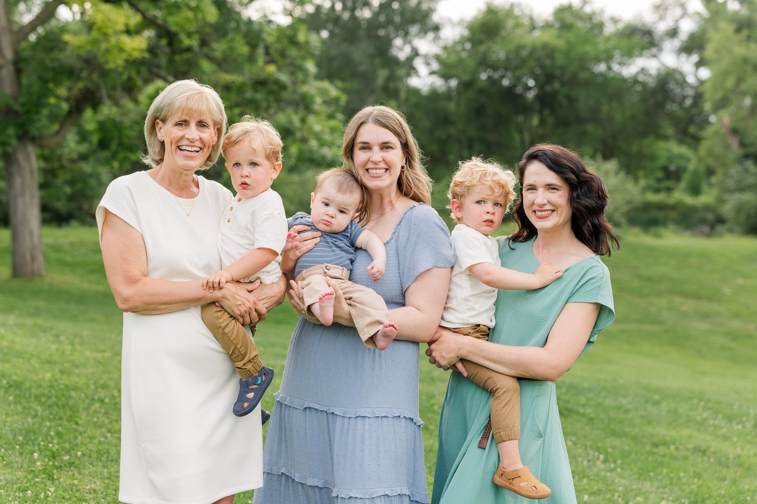 Three women smile outdoors in a green park, each holding a young toddler or baby in their arms.